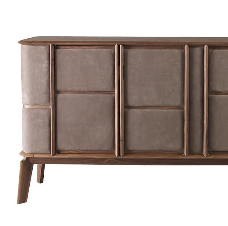 This elegant sideboard built in solid walnut features four short slanting legs that support a rectangular shape with characteristic curved corners highlighted by a series of front panels covered in leather that give its texture a dynamic and