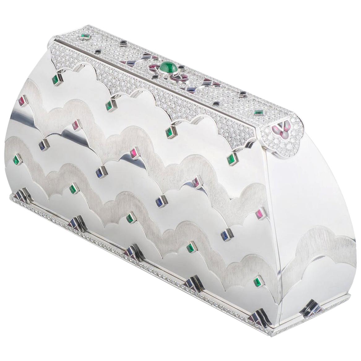 A unique 18k white gold Mauboussin multi-gemstone clutch bag. The clutch comprises of princess cabochon cut green tourmalines, sapphires and rubies placed evenly on the and back. There are diamonds set in a pave setting around the top and base of