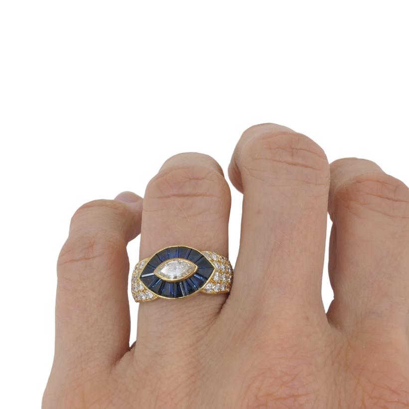 The ring features a striking eye-shaped motif that plays with symmetry and bold color. At the center is a marquise-cut diamond, bezel-set to emphasize its elongated shape. Surrounding it is a frame of deep blue baguette-cut sapphires, laid out in a