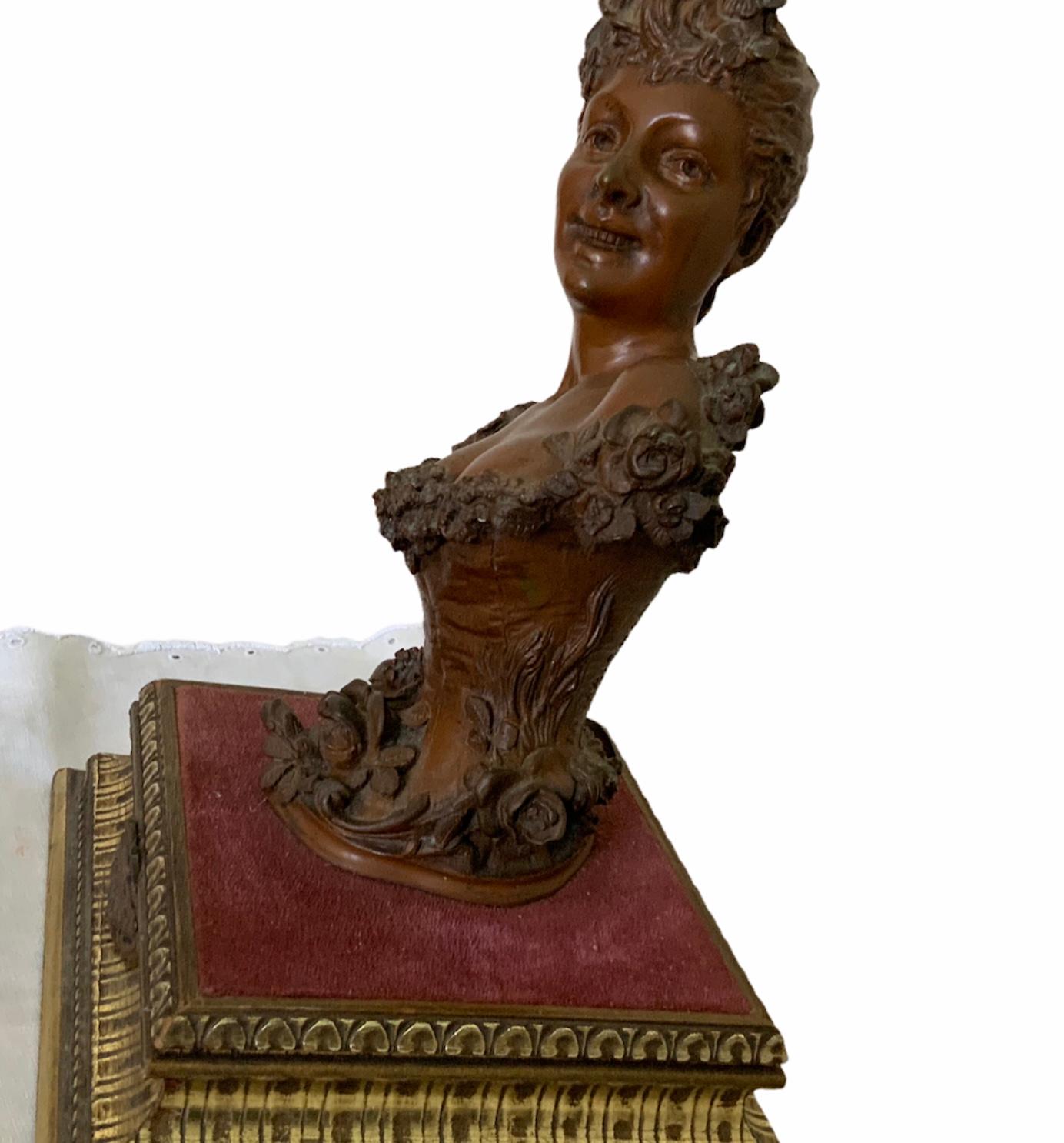 This is a Maurice Maignan bronze female bust sculpture. It depicts a young lady with a smile and wearing a tight corset adorned with an upper border full of ruffles. She also has like a garland of roses that start in her left shoulder and go around