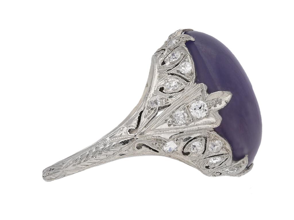 Mauve star sapphire and diamond cluster ring. Set to centre with an oval cabochon natural unenhanced mauve star sapphire with an approximate weight of 27.77 carats in an open back claw setting, further set with twenty three round eight cut diamonds
