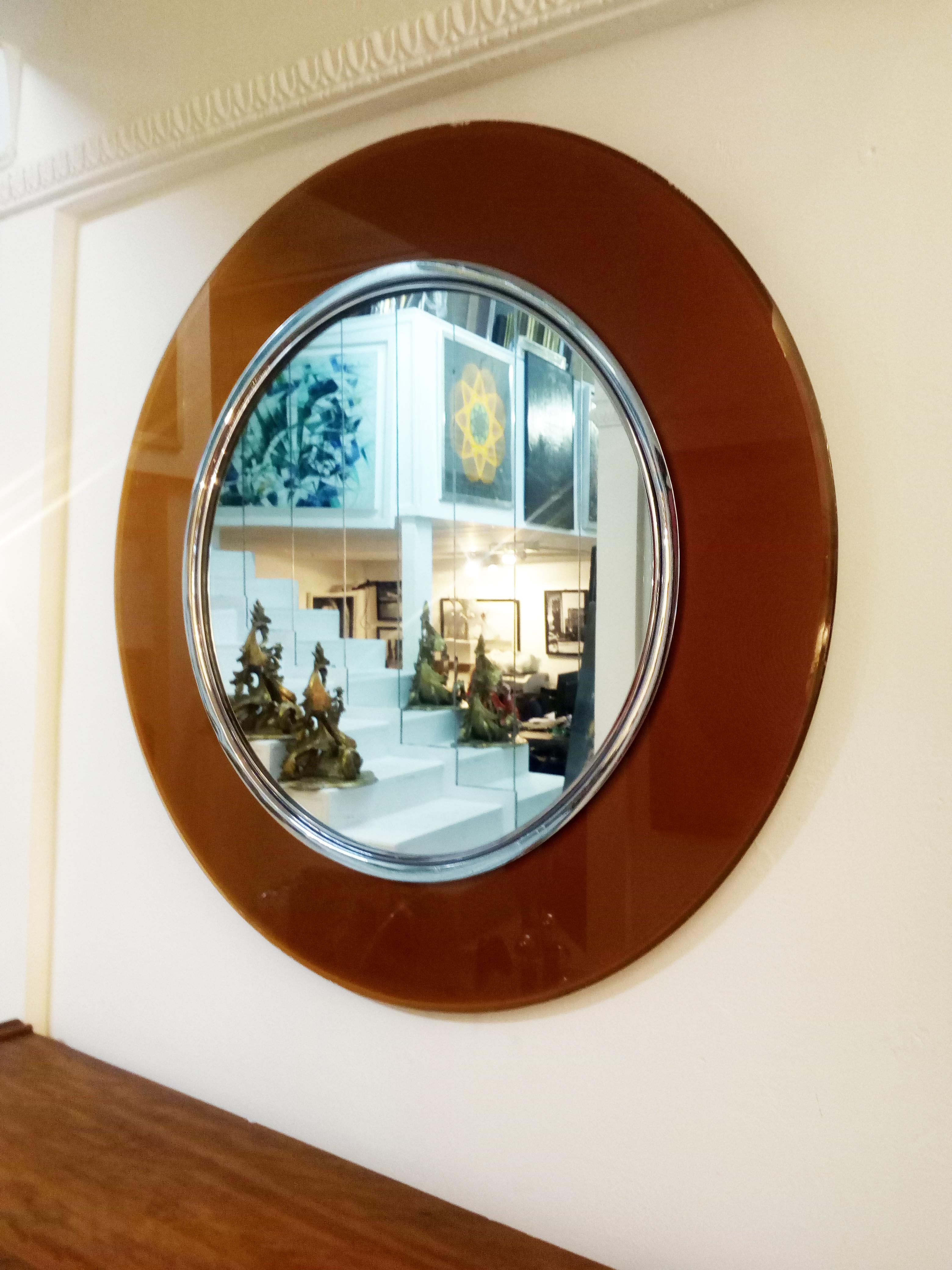 Colored, curved and beveled glass framing mirrored glass encased in brass trim.
This round mirror is created by Max Ingrand who works a lot with glass. This beautiful mirror is surrounded by a transparent silver amber color glass which is held