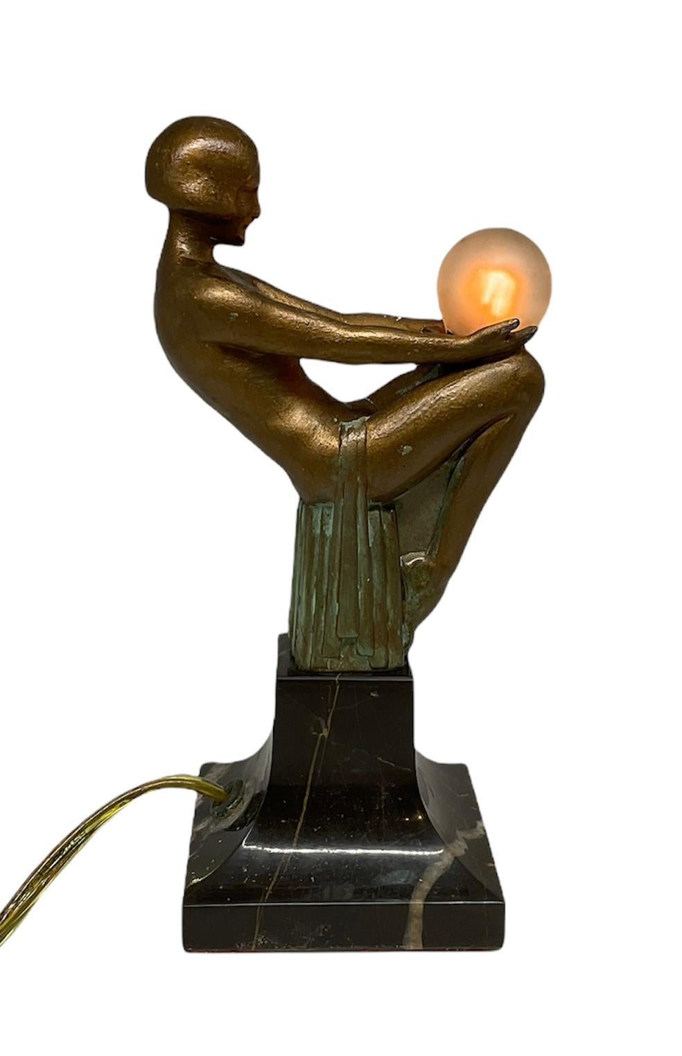 This is a Max Le Verrier “Songe Lumineux” Art Deco Lamp. It is depicting a 1920 bronze sculpture of a nude lady seated with her legs together in a rectangular block. A robe only covers her hip area while she is holding a light bulb. The sculpture is