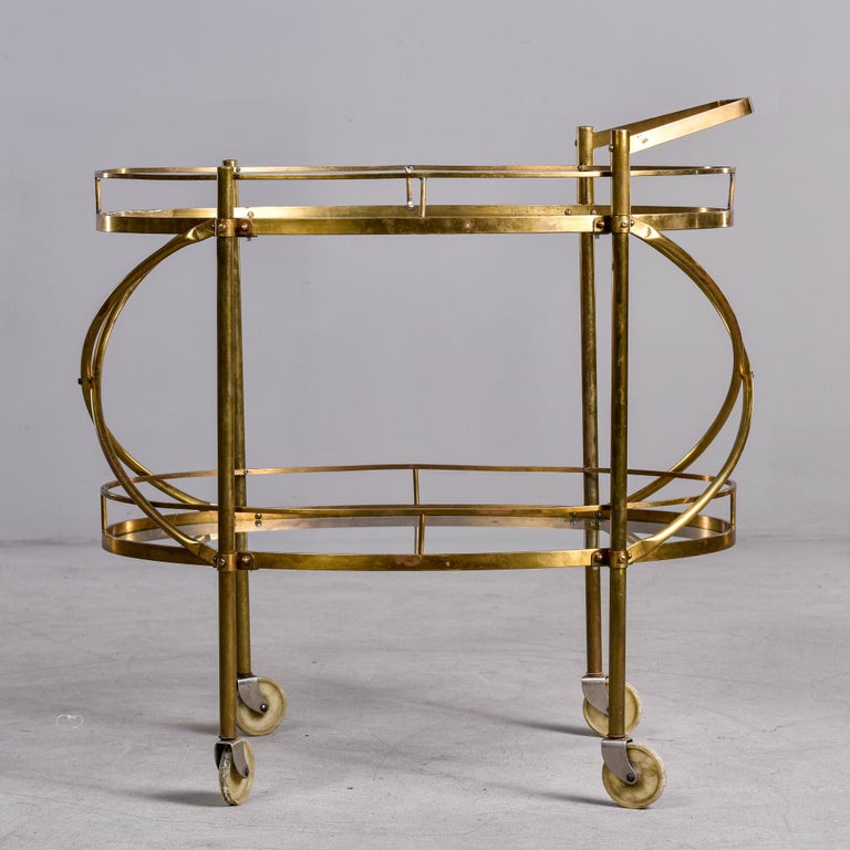 Found in the US, this circa 1960 two tier brass and glass bar cart is attributed to NYC retailer Maxwell Phillips. Each oval tier has a clear glass top with brass gallery. Top has a push handle and two tiers are connected at the ends with curved,