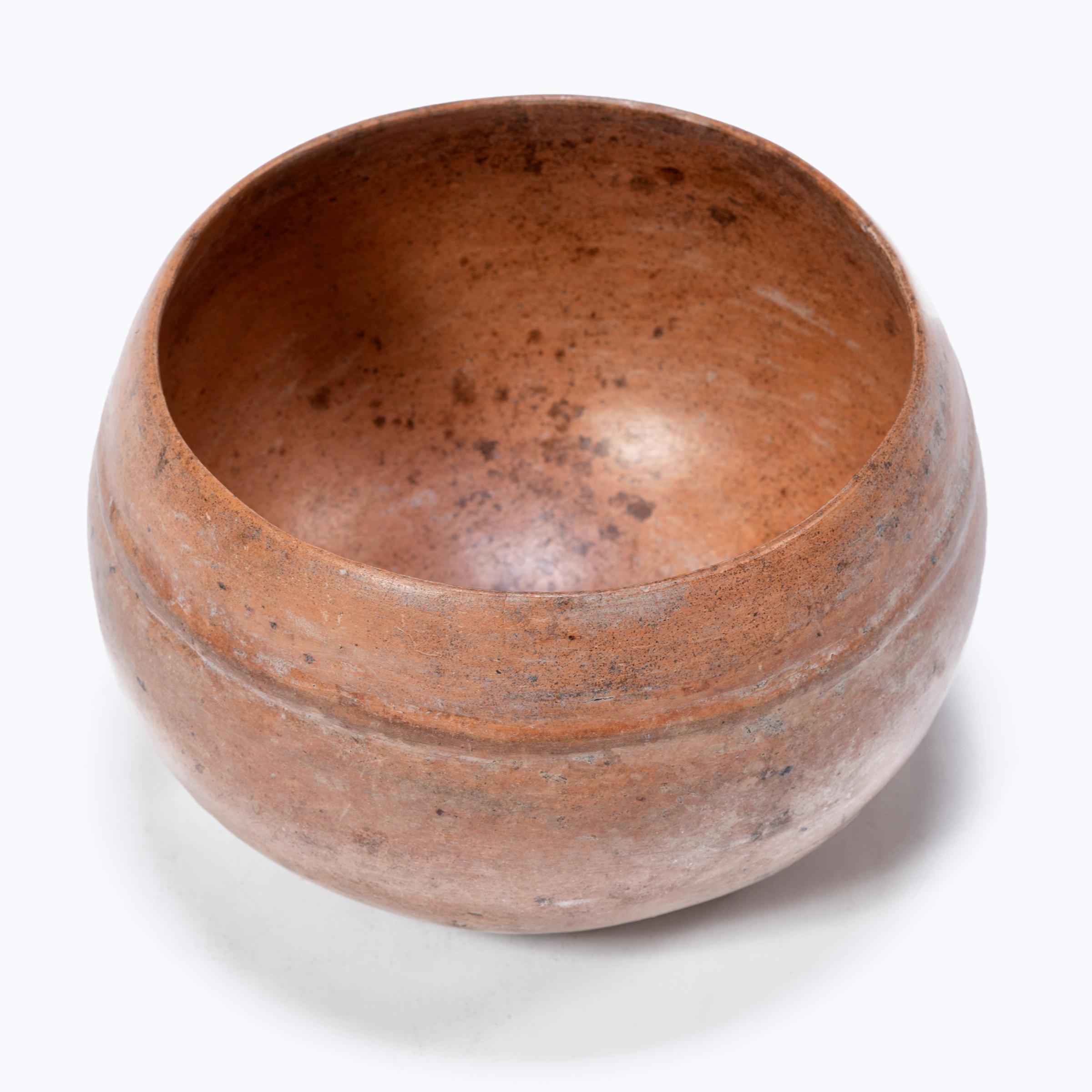 With a worn, glossy finish, this petite orangeware bowl is a wonderful example of Maya pottery. Coated in a layer of clay slip for a monochrome finish, the delicate bowl is bare of decoration save for a simple raised band along the lip. Centuries of