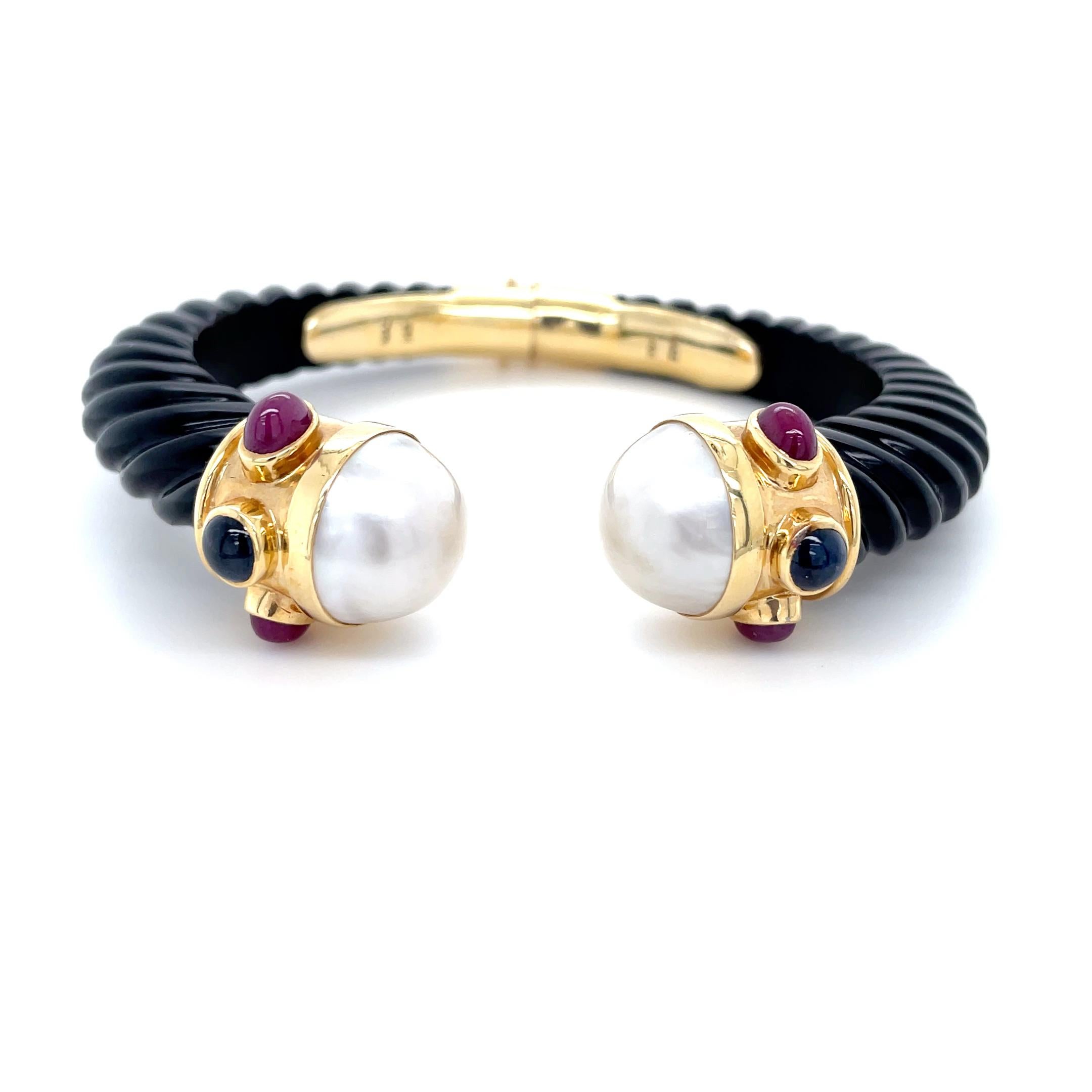This fun bangle by the designer Maz, features carved twisting black onyx with cultured mabe pearl terminals decorated with four bezel set oval cabochon rubies and two round cabochon sapphires. Mounted in 14 karat yellow gold and hinged at the back,