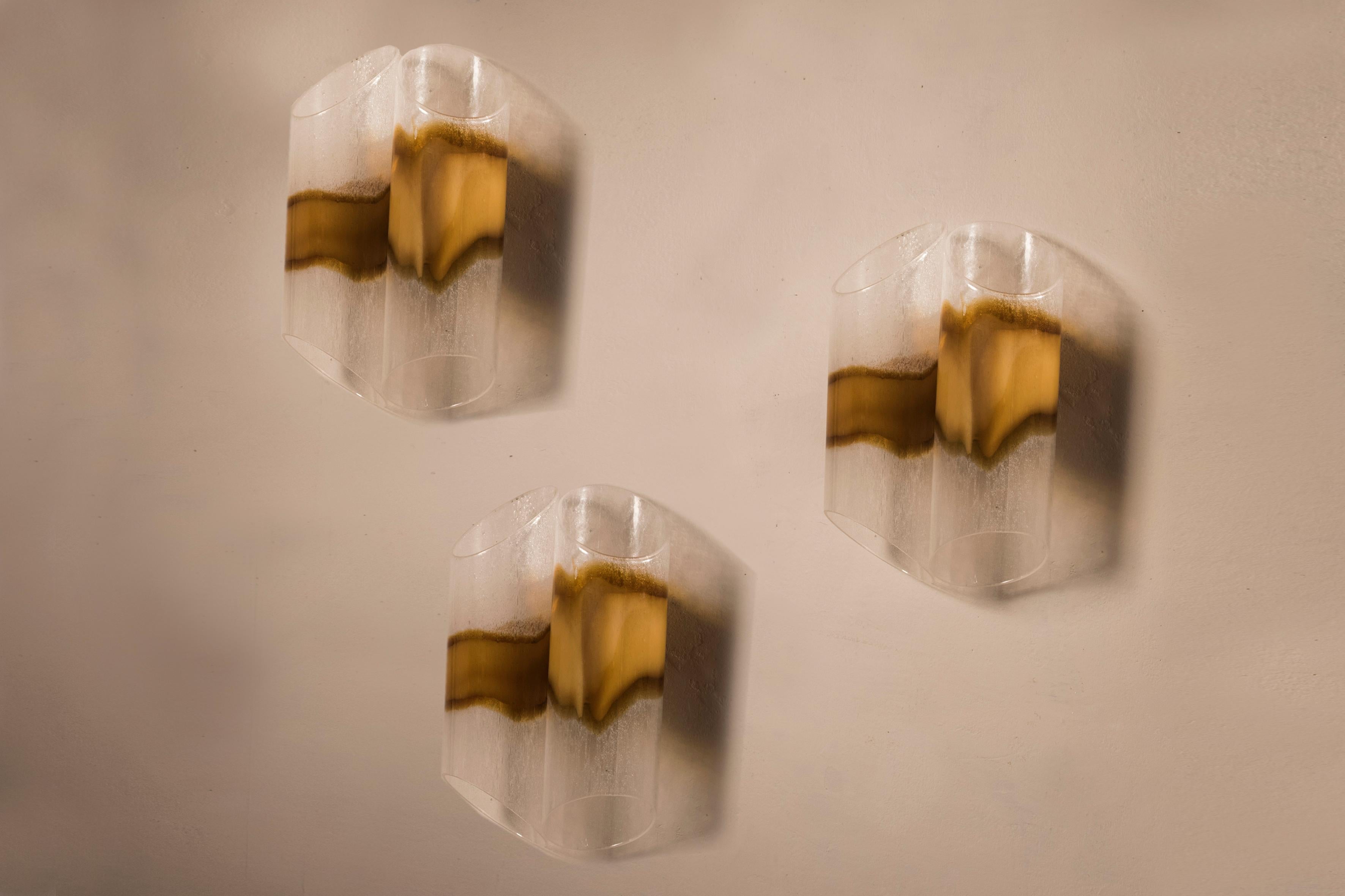 Set of three wall sconces by Mazzega. Italy, 1970s.
Each one is composed by two blown glass tubular parts mounted on a chromed steel base.
The glass shows different shades from transparent to ochre color. 
Each tubular part carries a light bulb