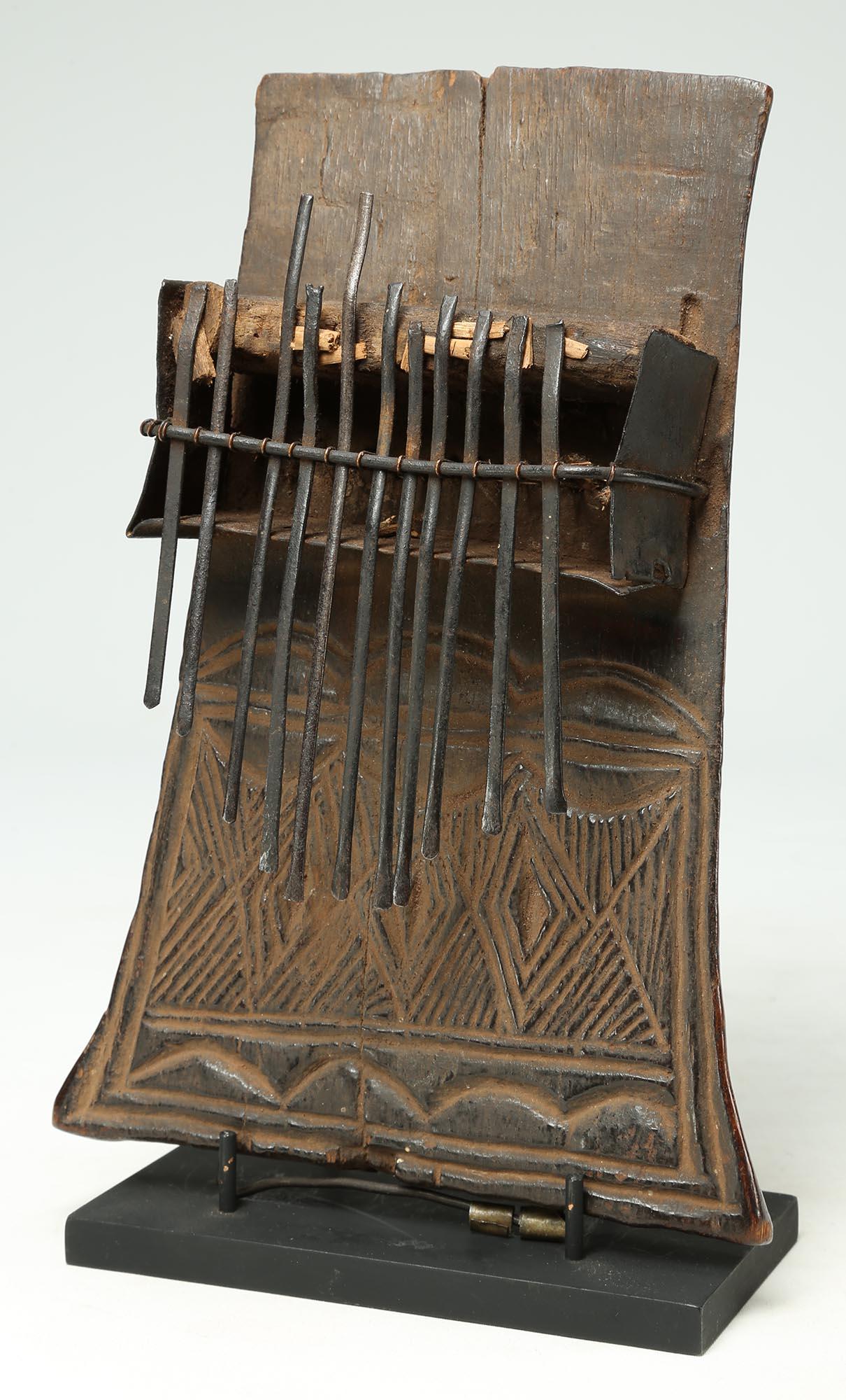 Mbira or Sanza (once known as a thumb piano), of carved wood with metal strikers. Under heavy patina is incised design with three stylized cowrie shells (also resemble human eyes) over a fluid geometric pattern. Created in the Congo region in the