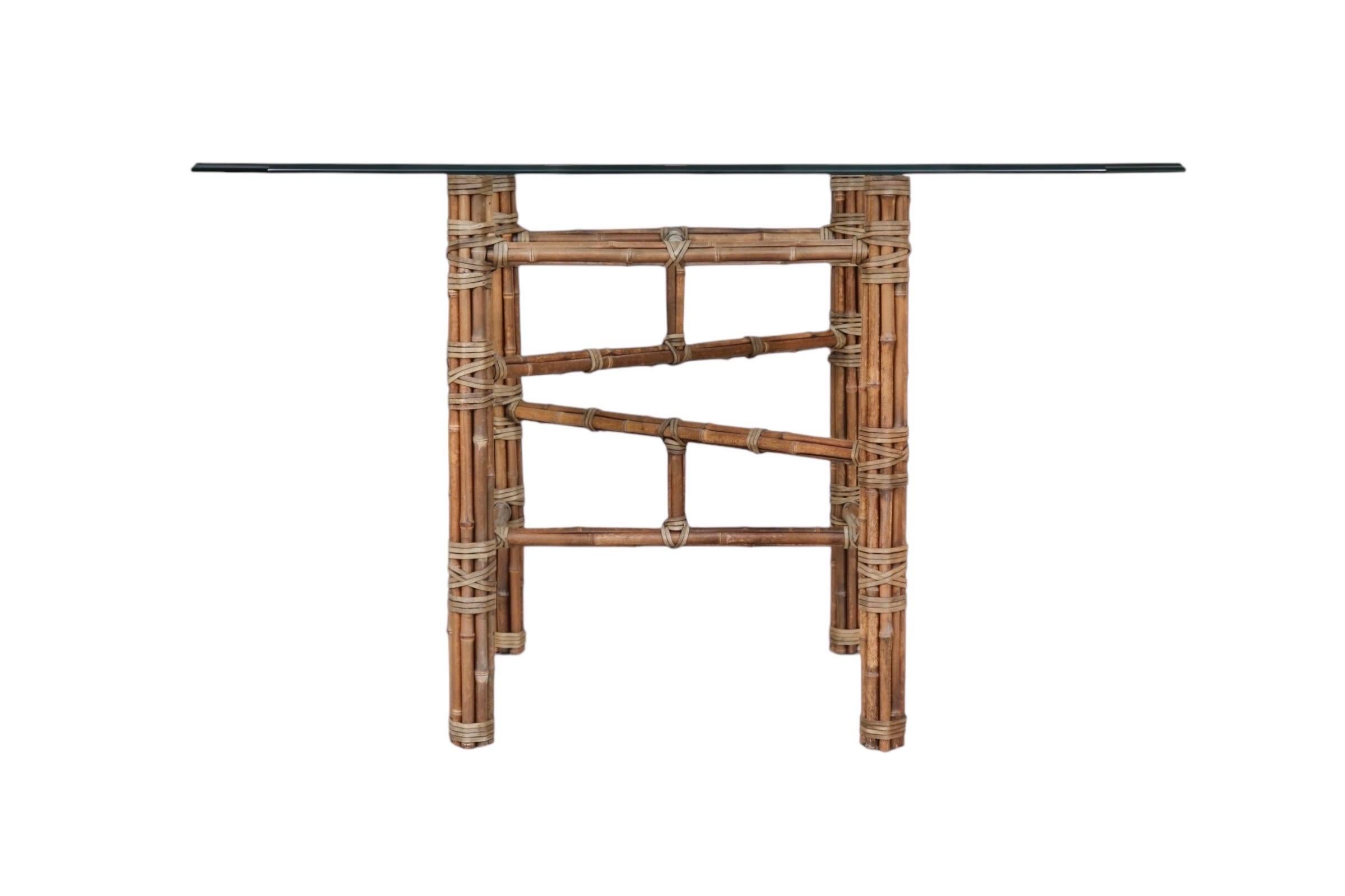 An organic modern bamboo rattan and glass dining table attributed to McGuire. The thick glass tabletop has a curved beveled edge and cut corners. Below, the table base consists of four legs and two H-stretchers with additional cross stretchers and