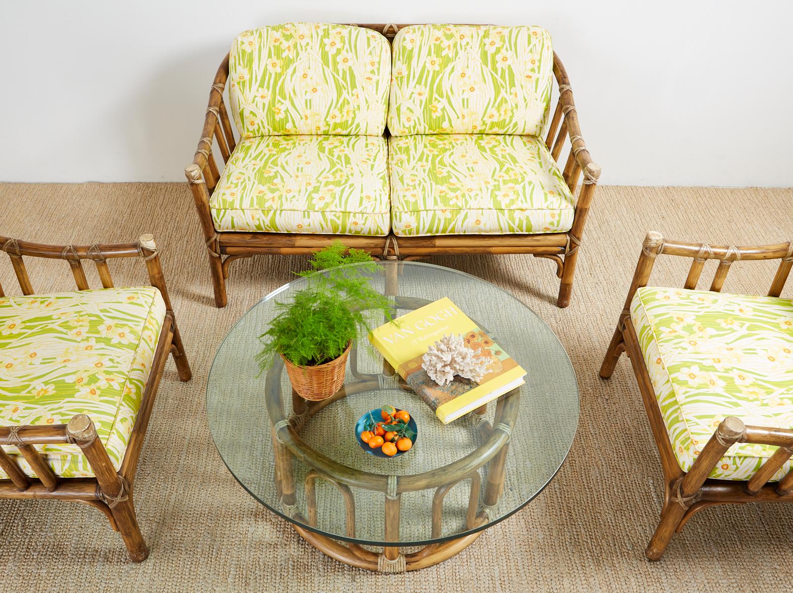 California organic modern style living room suite by McGuire. Set consists of a settee, two lounge chairs, and a round coffee cocktail table. Made from rattan poles lashed together with leather rawhide laces. Topped with bright floral motif seat