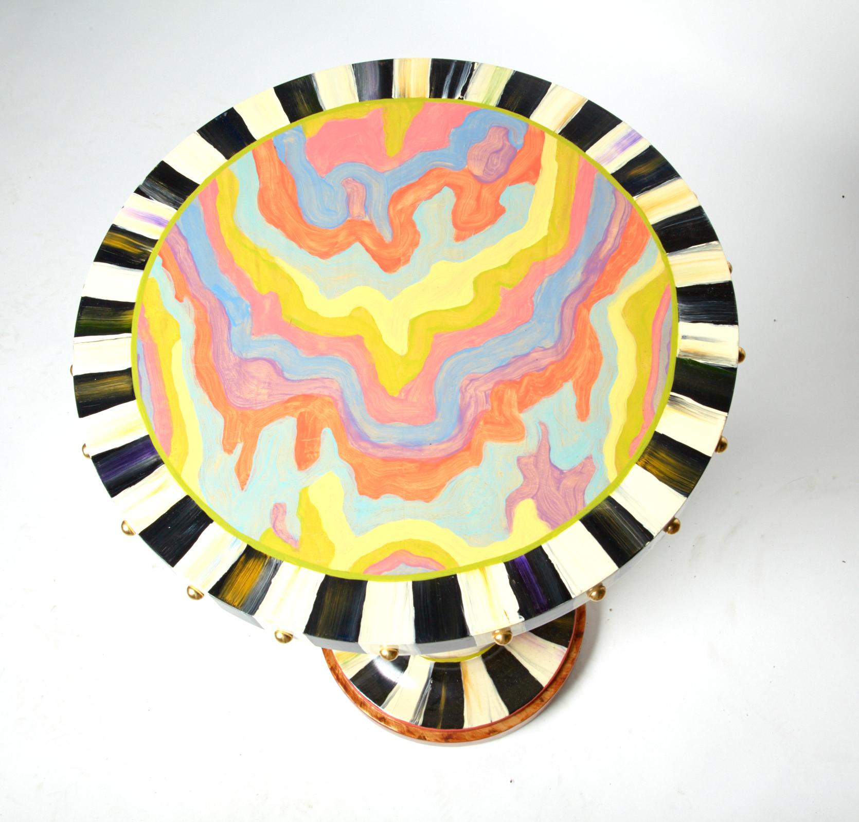 Colorful MacKenzie-Childs pedestal table in black and white with a colorful swirled painted top. The rim is decorated with brass domed tacks. 2 of them are dimpled. Otherwise the table is in excellent condition and the colors are bright. it has the