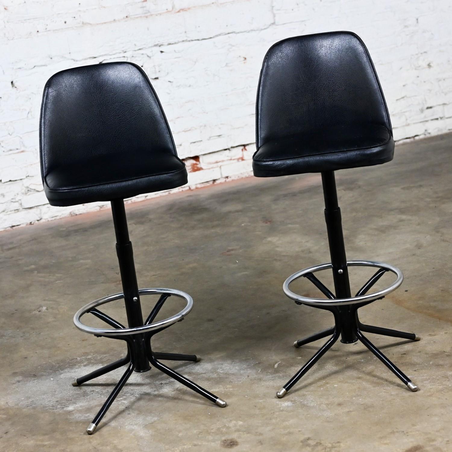 Handsome vintage MCM or Mid-Century Modern adjustable barstools comprised of black vinyl or faux leather bucket seats, black painted steel manually adjustable shaft, chrome footrest, and black painted steel base composed of four legs with chrome