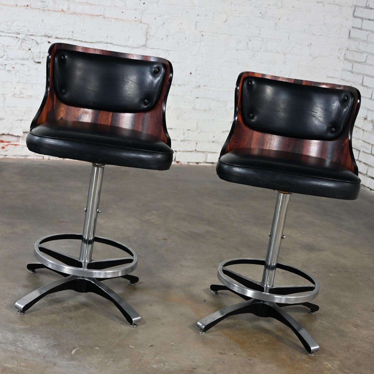 Handsome vintage Mid Century Modern adjustable and swivel barstools comprised of the original black vinyl or faux leather, molded, curved faux rosewood laminate backs, chrome shaft, and cast aluminum four prong base & footrest. Beautiful condition,