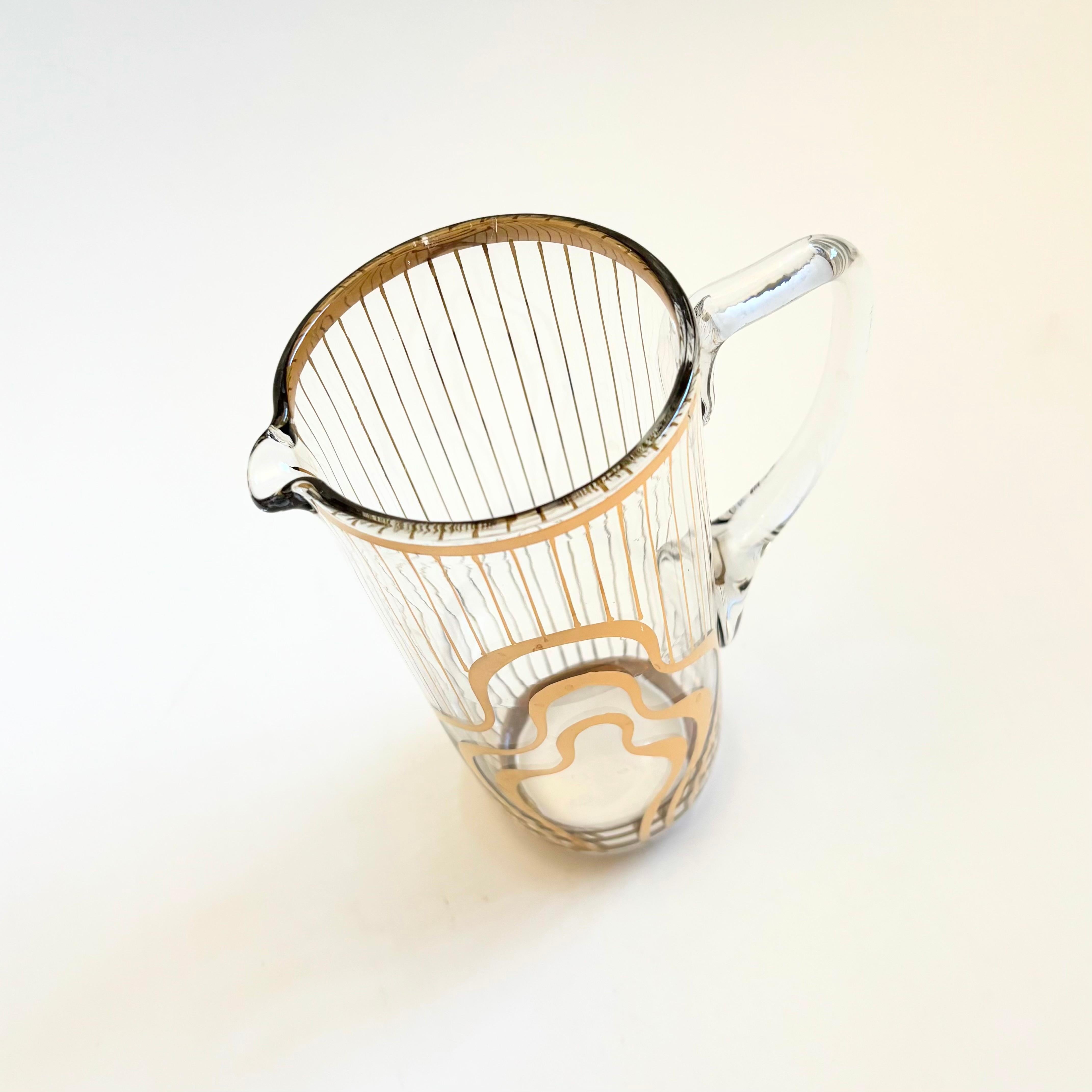 A mid century glass pitcher decorated in a metallic gold squiggle and stripe design.

