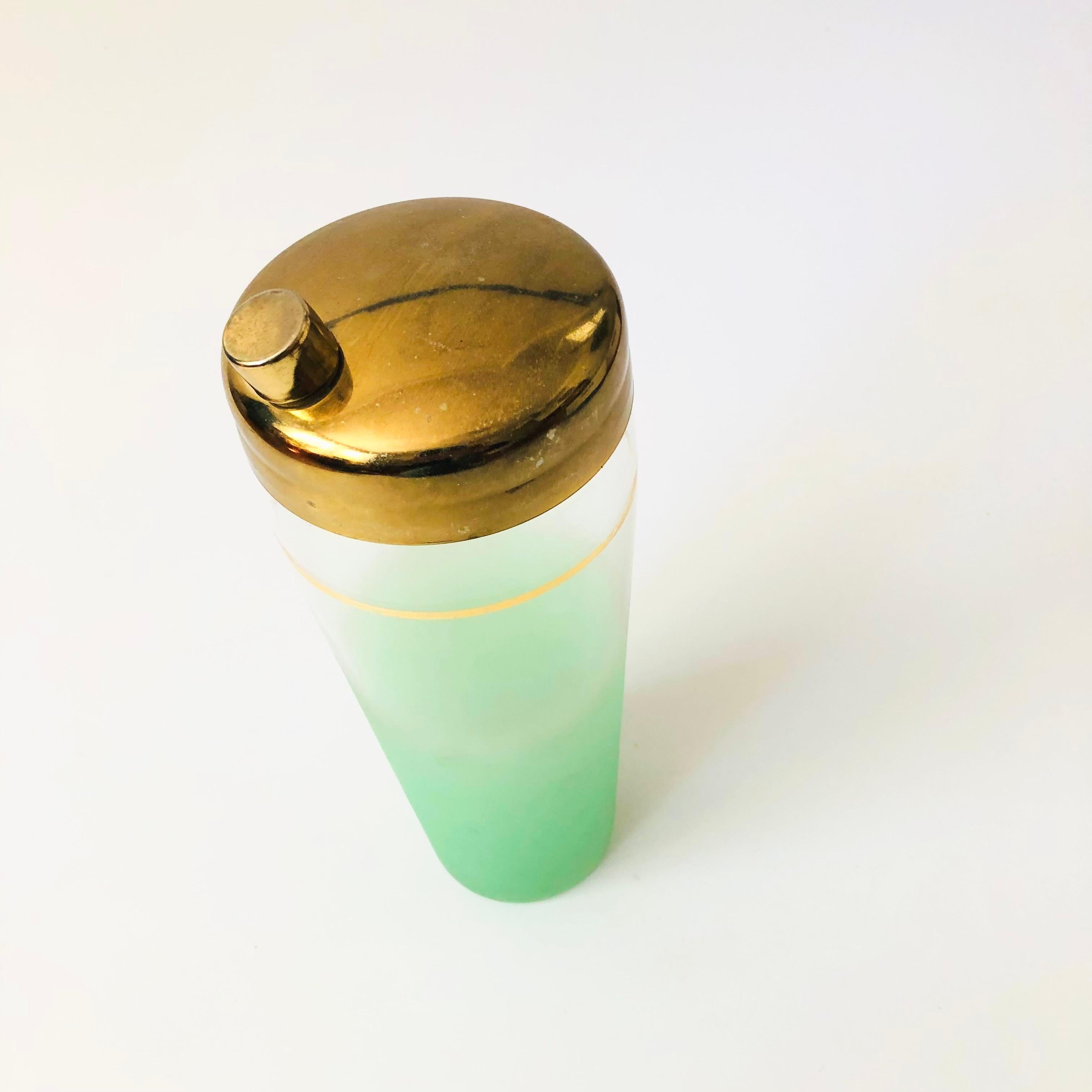 A  mid century glass cocktail shaker. Beautiful green blendo coloring to the glass the fades from bright green at the base to clear at the top. A band of metallic gold decorates the upper part of the glass. Topped off with a gold tone metal cap.



