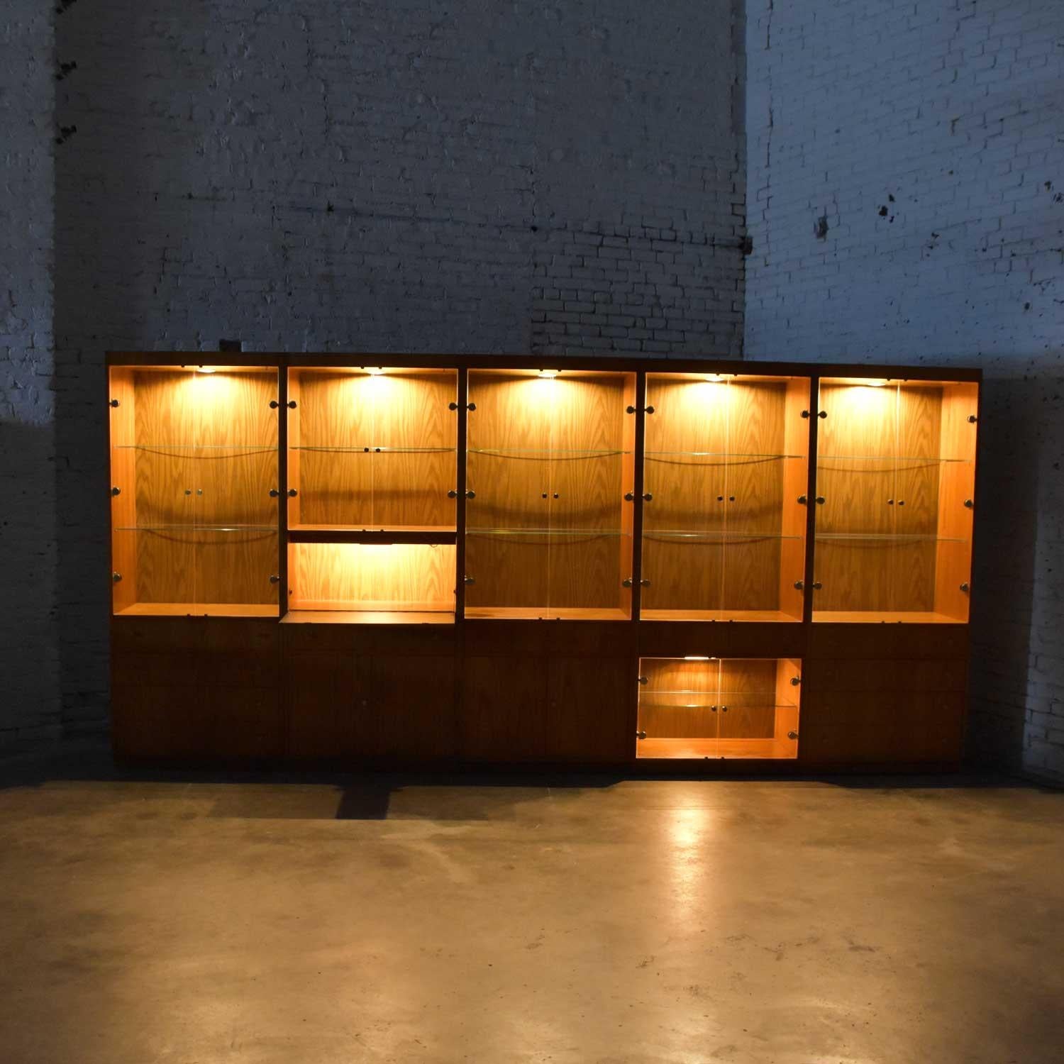 Gorgeous Mid-Century Modern (MCM) five section oak veneer lighted display cabinet wall unit with smoked glass doors, brass pulls, ten glass shelves with plate grooves, two wooden shelves, and brass plate hinge covers by Hooker. Beautiful vintage