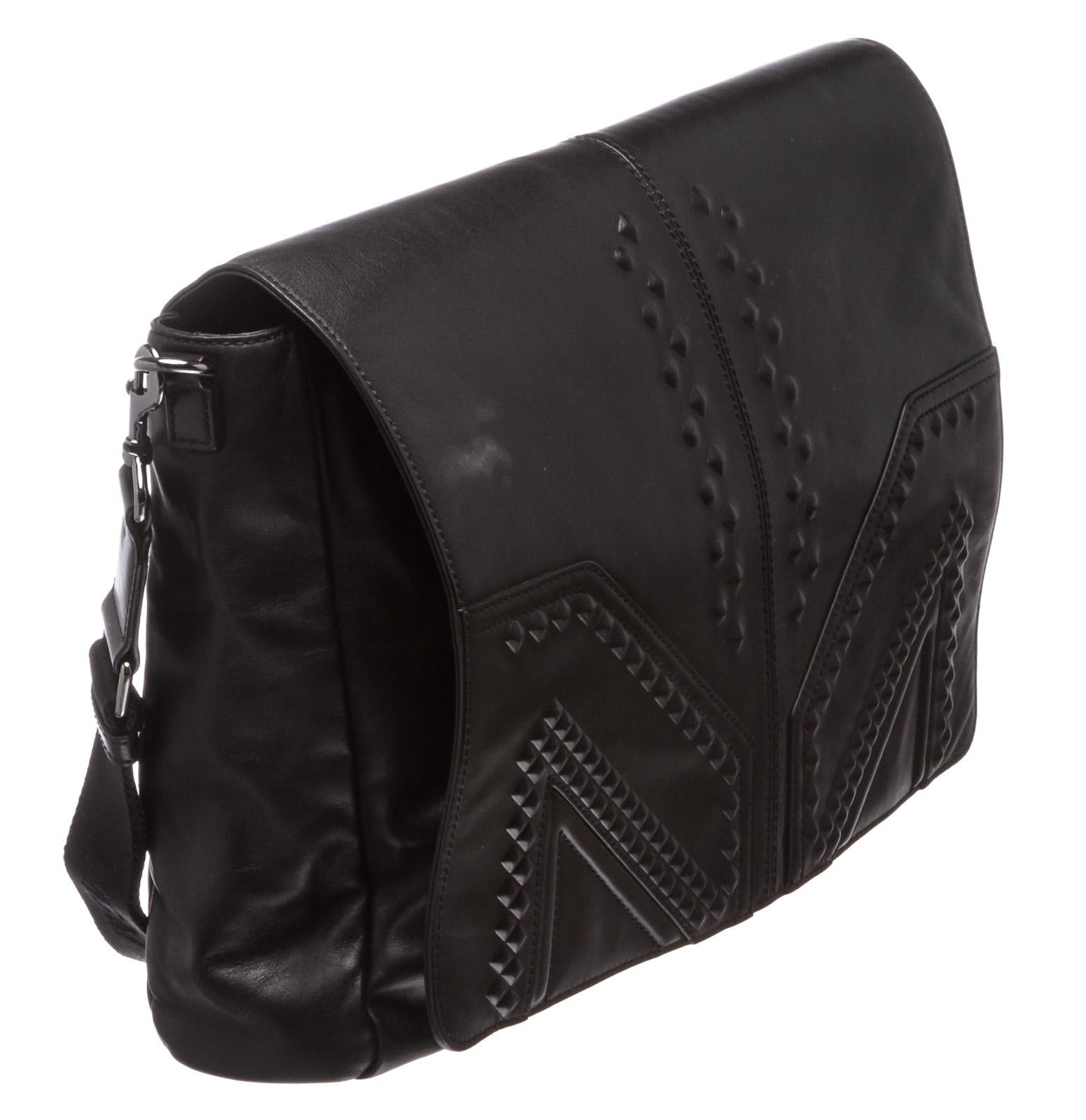 Black leather MCM M. Moment messenger bag contains tonal stitching, debossed stud details at front face and detachable shoulder strap. Interior is satin lined and contains two compartments, one zipper two zipper pockets, and two side slip pockets.