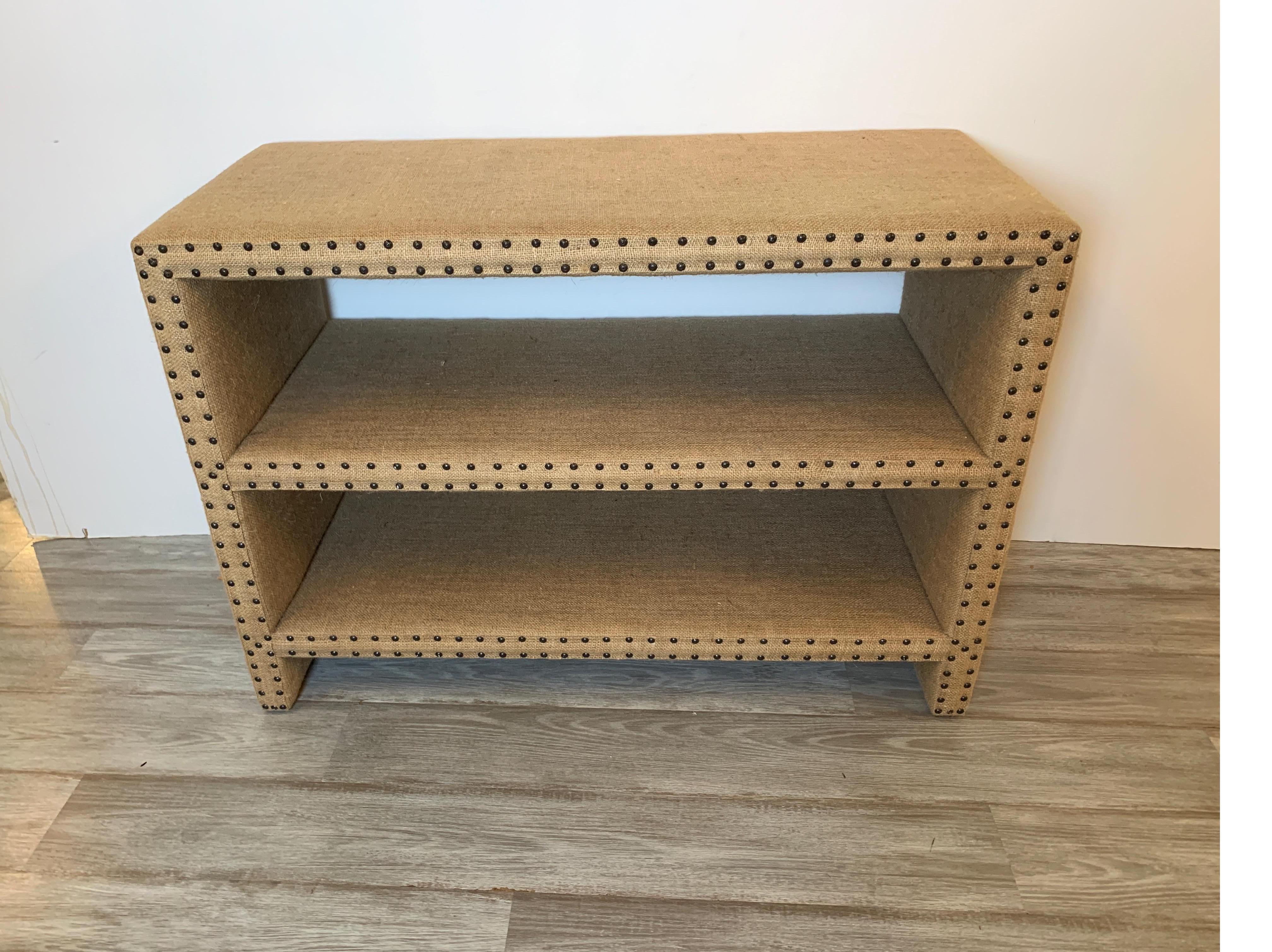 Mecox Parsons style bookshelf or side table upholstered in burlap with nailhead trim.