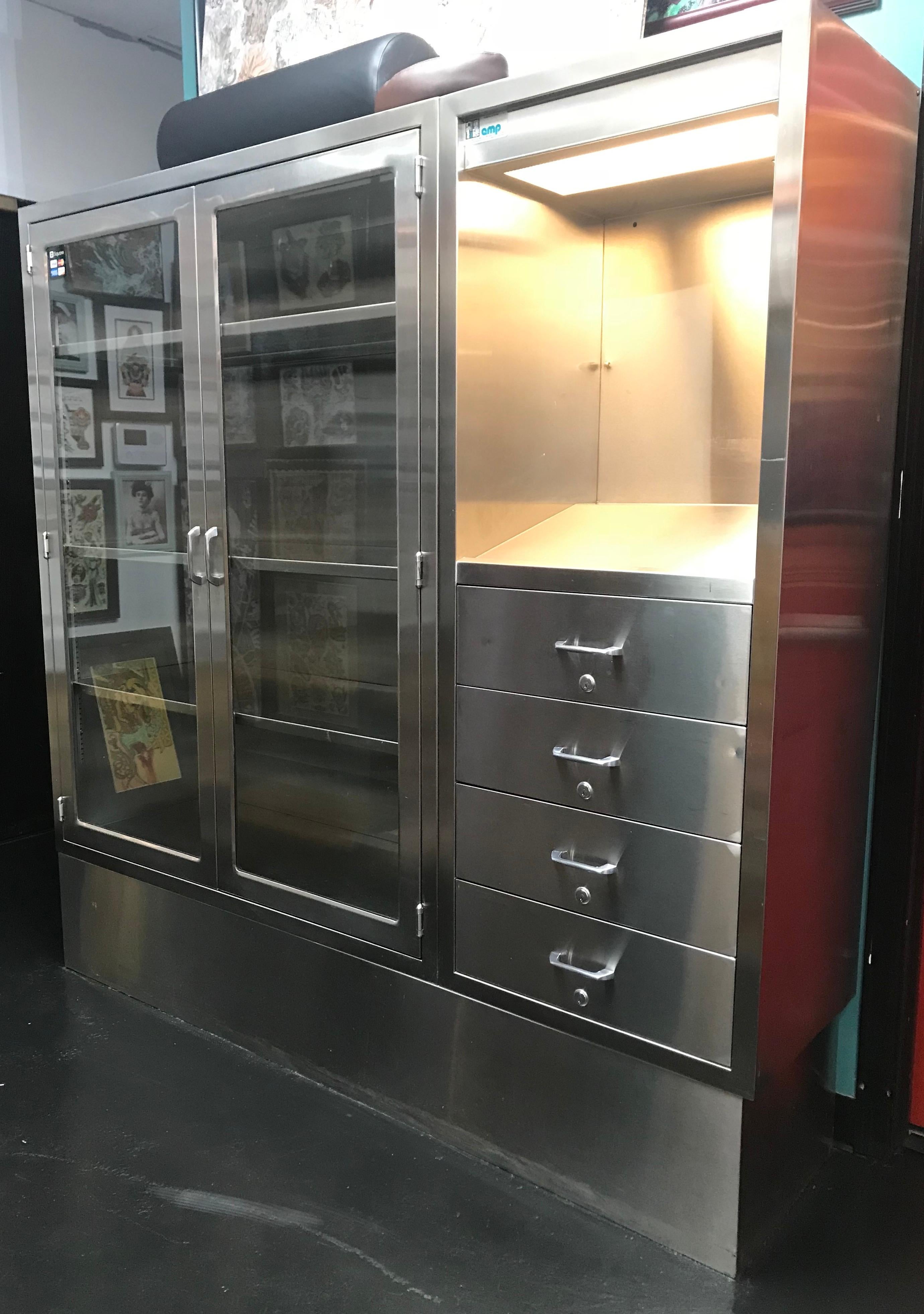 Stainless steel medical cabinet with four adjustable shelves and tempered glass door panels. Has working lighting display and four locking drawers. Extensive and secure storage capacity. Very sharp. Comprehensive. A significant piece. Two identical