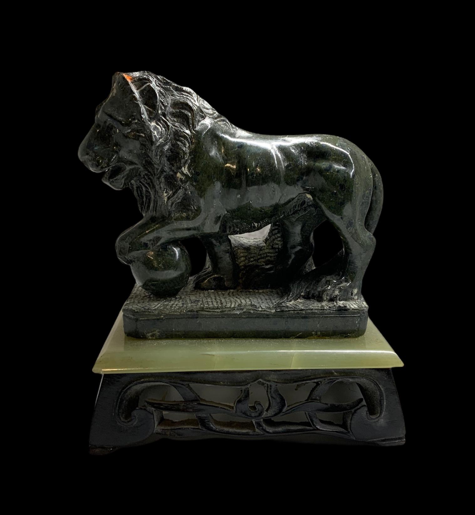 This a carved sculpture of what appears to be granite stone of a lion holding a ball with his paw. It is standing over a rectangular light green onyx plaque that is supported by a dark wood rectangular pierced base adorned with branches of leaves.
