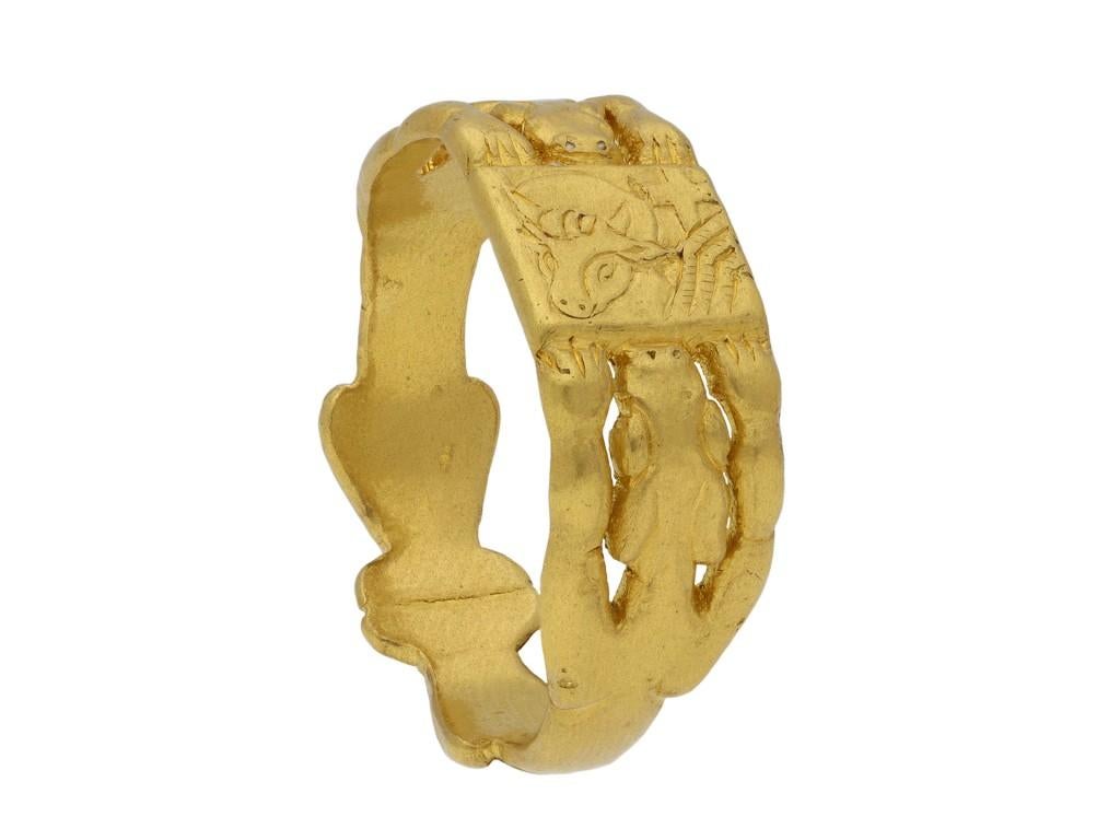 A yellow gold ring with central rectangular plaque finely engraved with an ox head with halo, wings and cross, the flat shank composed of a mirrored pair of outstretched finely carved openwork beasts, with forelegs supporting the plaque, the