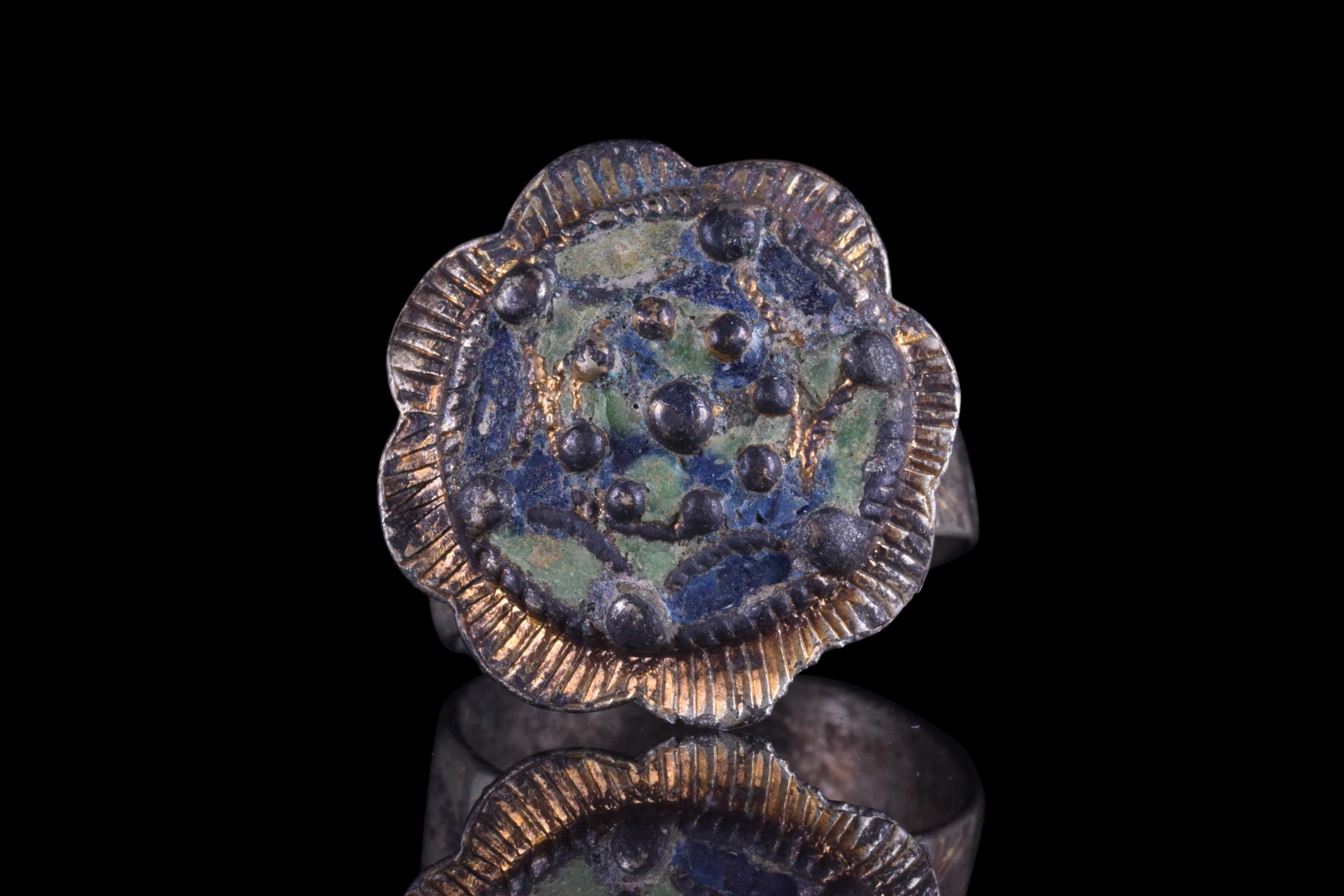 An exquisite silver gilt finger ring featuring a beautiful floral-shaped bezel with raised bosses and remnants of blue and green enamel. The ring is further embellished with several intricate details, including delicate engravings on the stepped