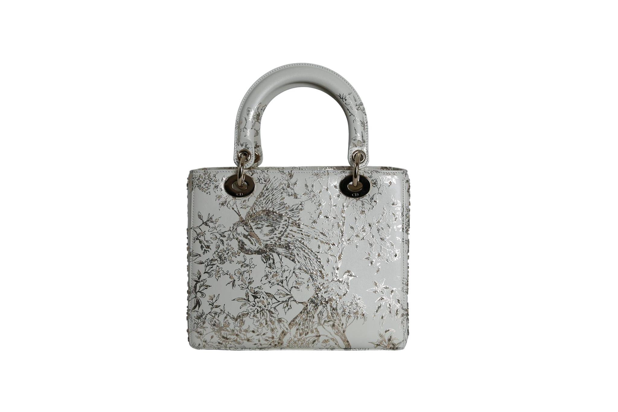 Medium Lady Dior bag in smooth calfskin adorned with golden embroidery in a white and gold/silver design. With its versatile design, this bag is ideal for a variety of occasions, adding a touch of sophistication to any outfit.

Logo charms to the