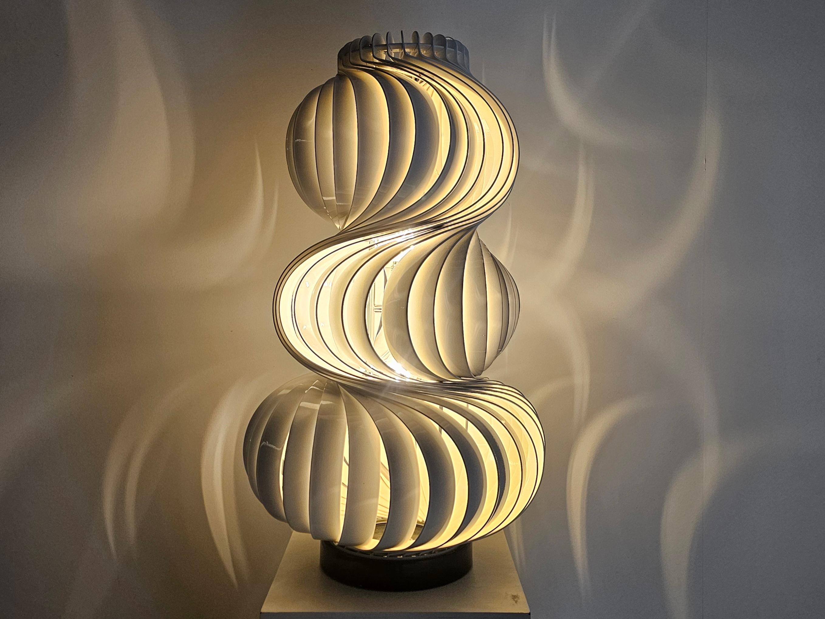 The ‘Medusa’ features a round black metal base and a central chrome spine that supports both the 2 sockets and the outer shade with an ingenious mechanism. The shade is composed of several bent strips of offwhite-finished aluminum, artfully rotated