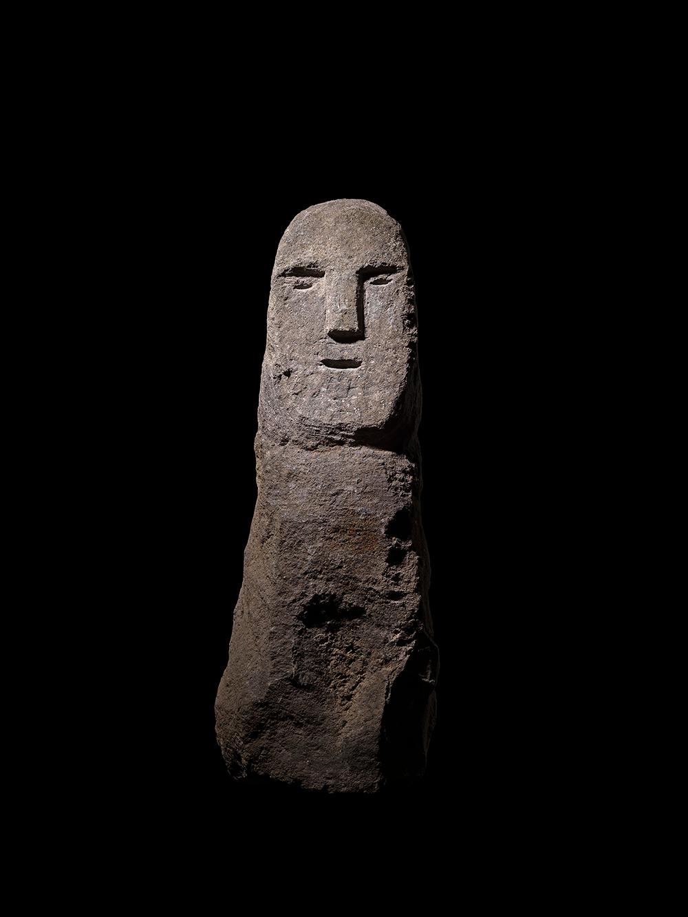 A tall anthropomorphic stele of carved granite, divided into two distinct regions of the body and face. The body is a single unarticulated block, but the facial features are outlined with strong carved lines. A T-shape marks the position of the nose
