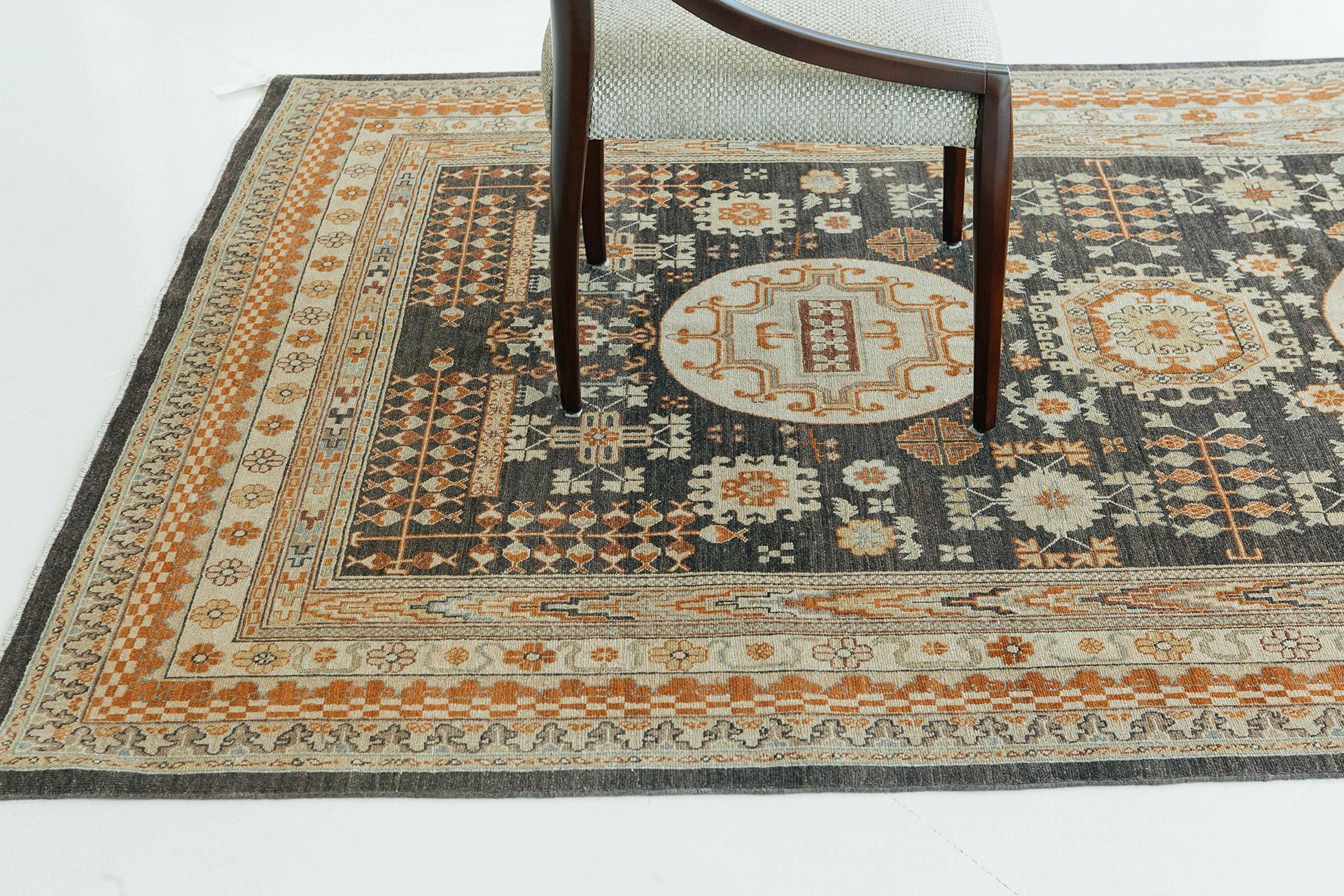 Un tappeto in stile Khotan annodato a mano e realizzato in lana, ha un design, motivi e bordi distinti. Lo sfondo antracite scuro, insieme ai toni del ruggine, del marrone van dyke e del caramello, crea una decorazione affascinante e versatile per
