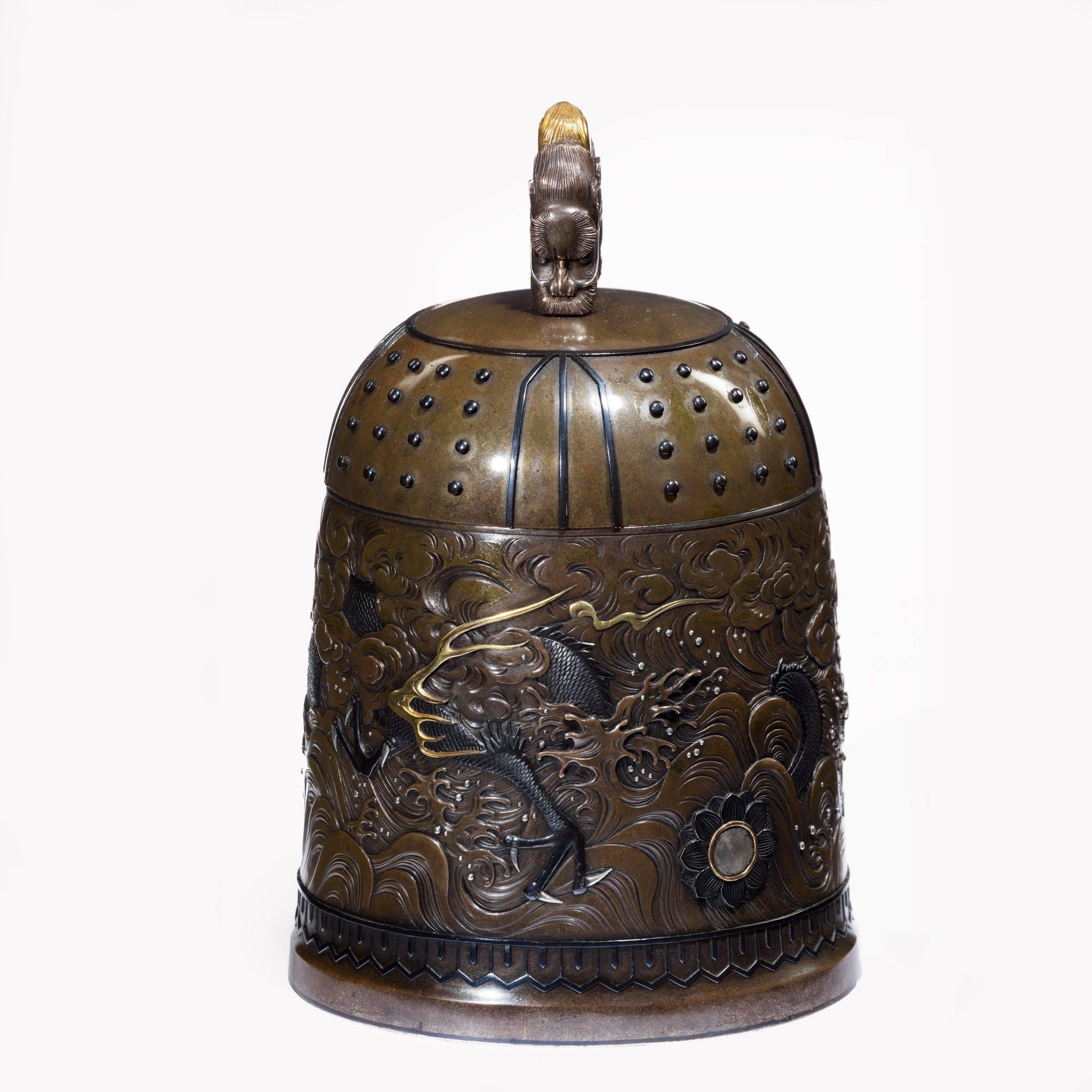Eine hervorragende Glockenschatulle aus gemischtem Metall aus der Meiji-Zeit von der Gießerei Nogowa,
Typische Form mit Shibuichi-Grund, gearbeitet in Shakudo, Silber und Gold, mit einem durchgehenden Fries in der Mitte, der einen dreizackigen