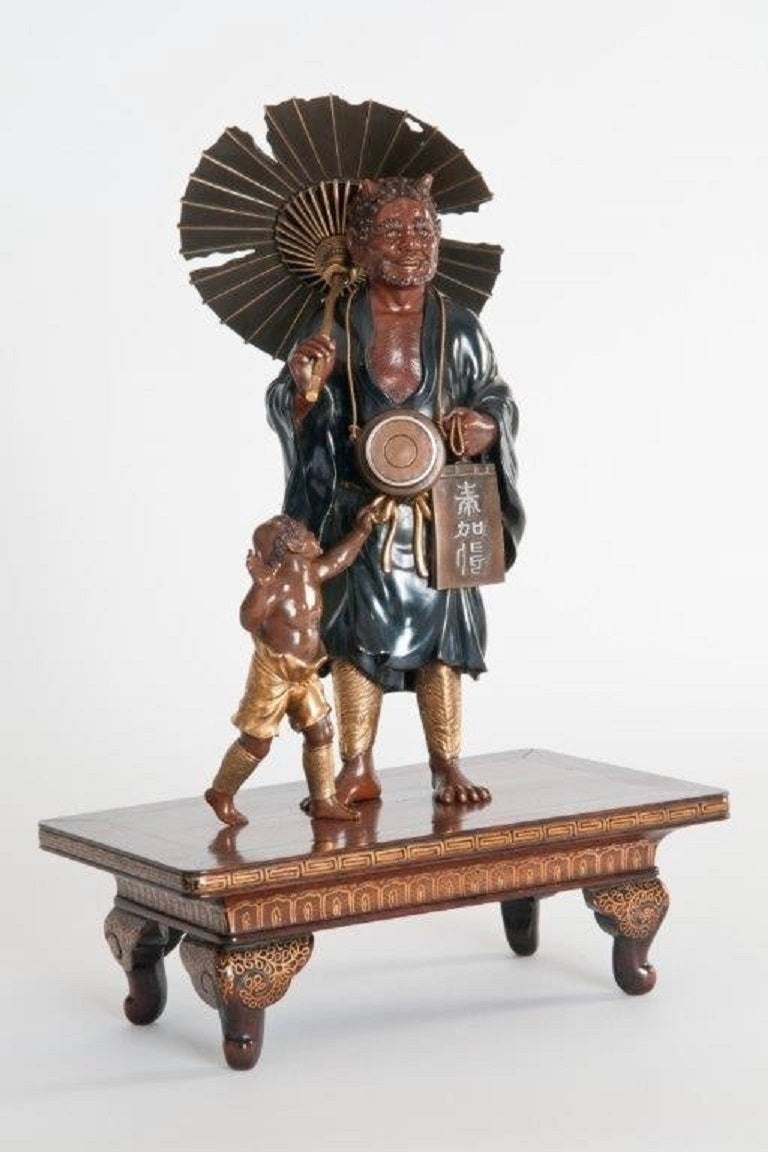 This figural group shows a mendicant Oni with a tattered umbrella and wearing a voluminous shakudo cloak over gilt tiger mask greaves. A smaller Oni wears similar greaves and a gilt loin-cloth. Set on a gilt decorated wood stand. Meiji period.