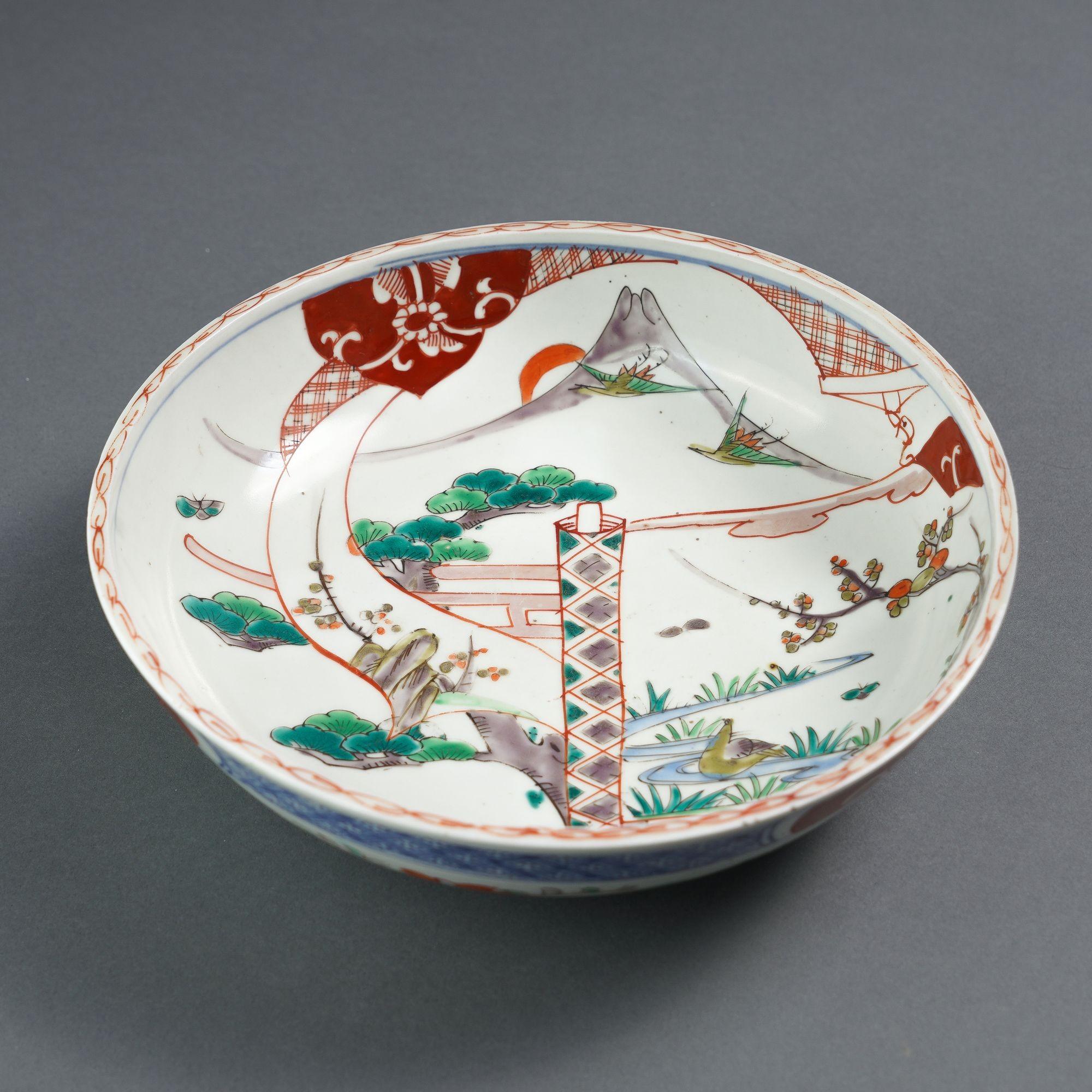 Imari palette shallow bowl decorated in Imari colors simulating the Chinese famille verte palette (green with accents of aubergine, oxide red and underglaze blue). The interior of the bowl is decorated with segmented images of mountain, a stream,