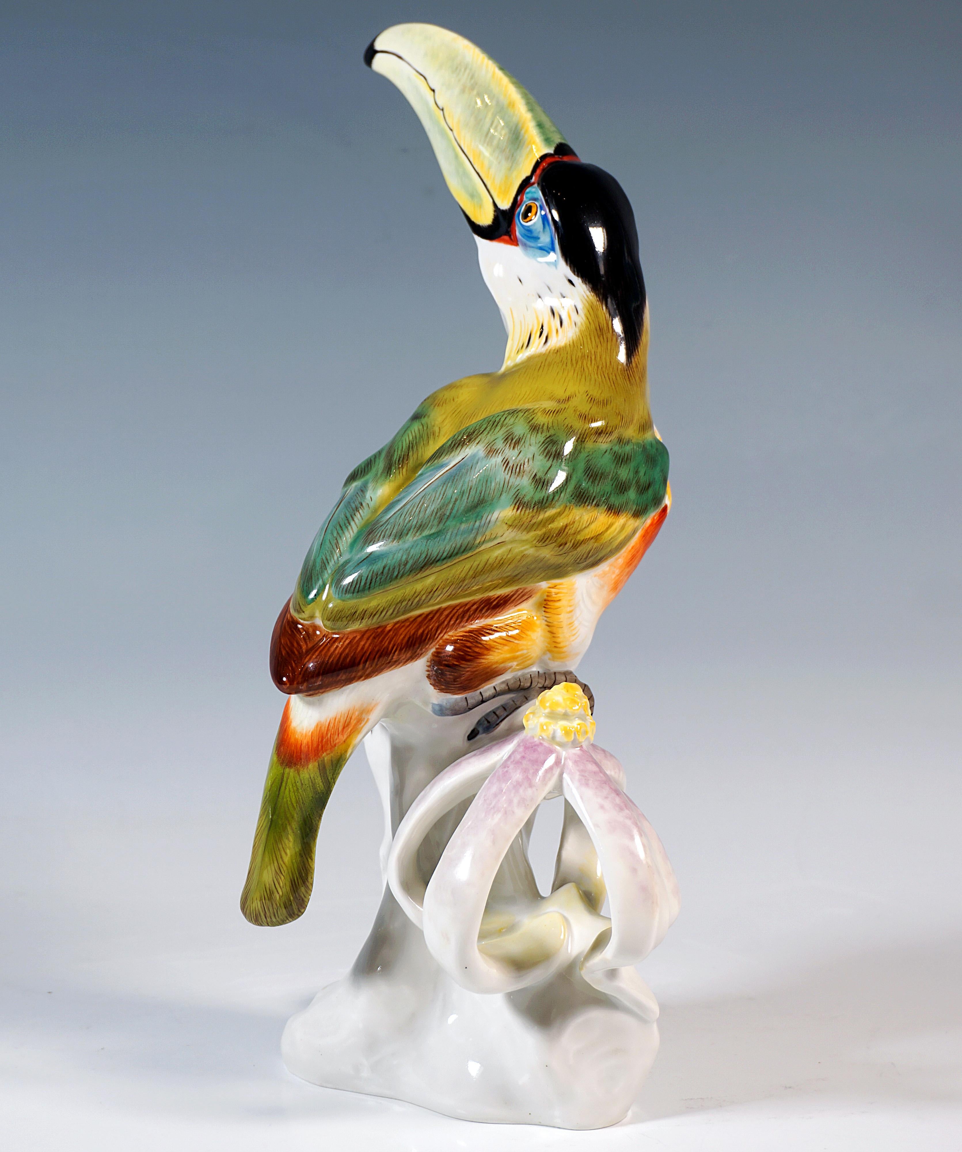Highly decorative depiction of a life-size toucan clinging to a tree trunk with its beak raised to the left.
Colourfully painted, on an elongated white base with a rooted tree stump, to the right of the bird is a plant reminiscent of a sun