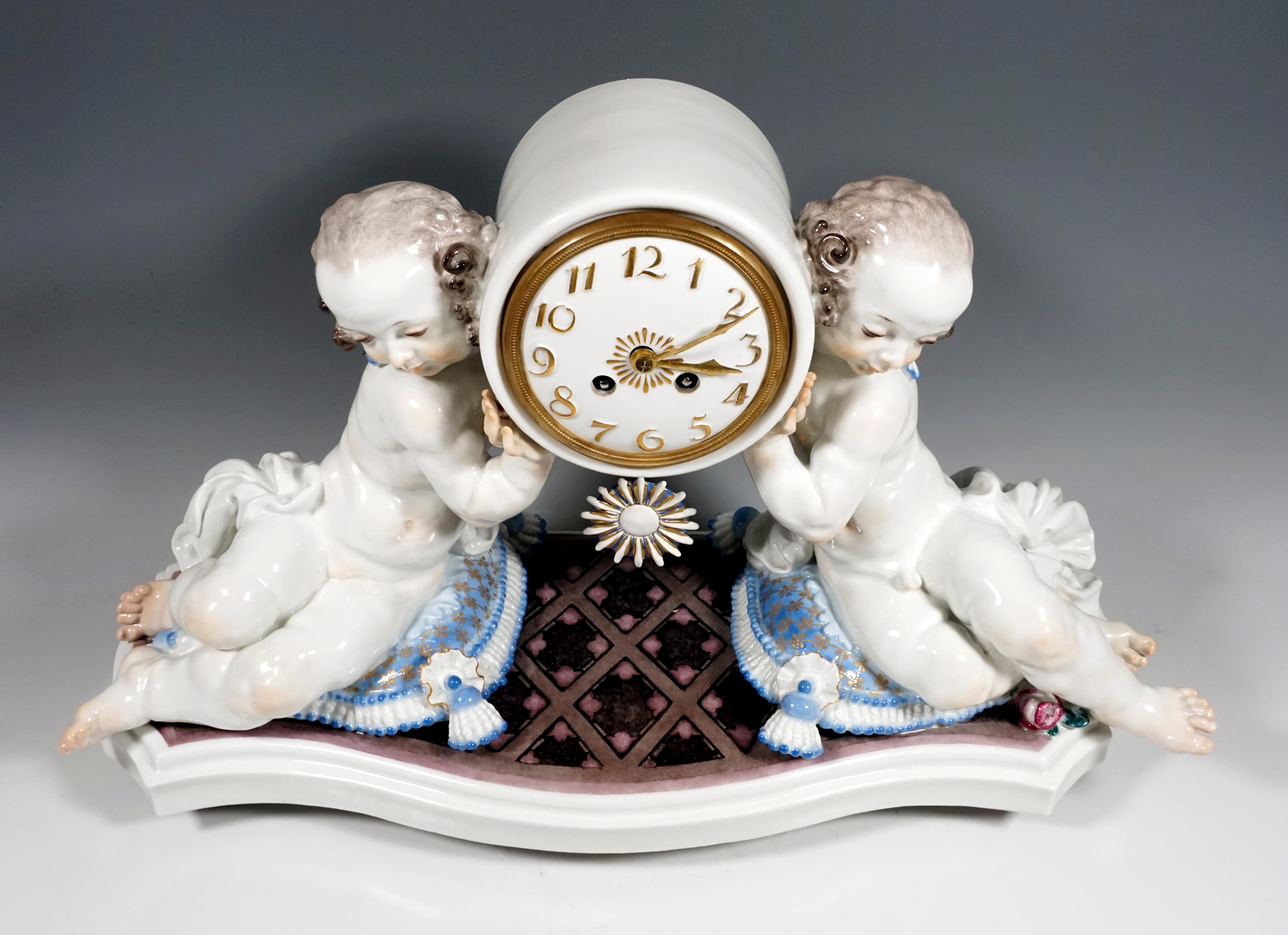 On a curved base plate with diamond pattern in marble look, sculptured cushions with tassel decoration, on the side seated figures, girl and boy, who gracefully carry the round clock case. White porcelain dial with applied Arabic numerals and hands