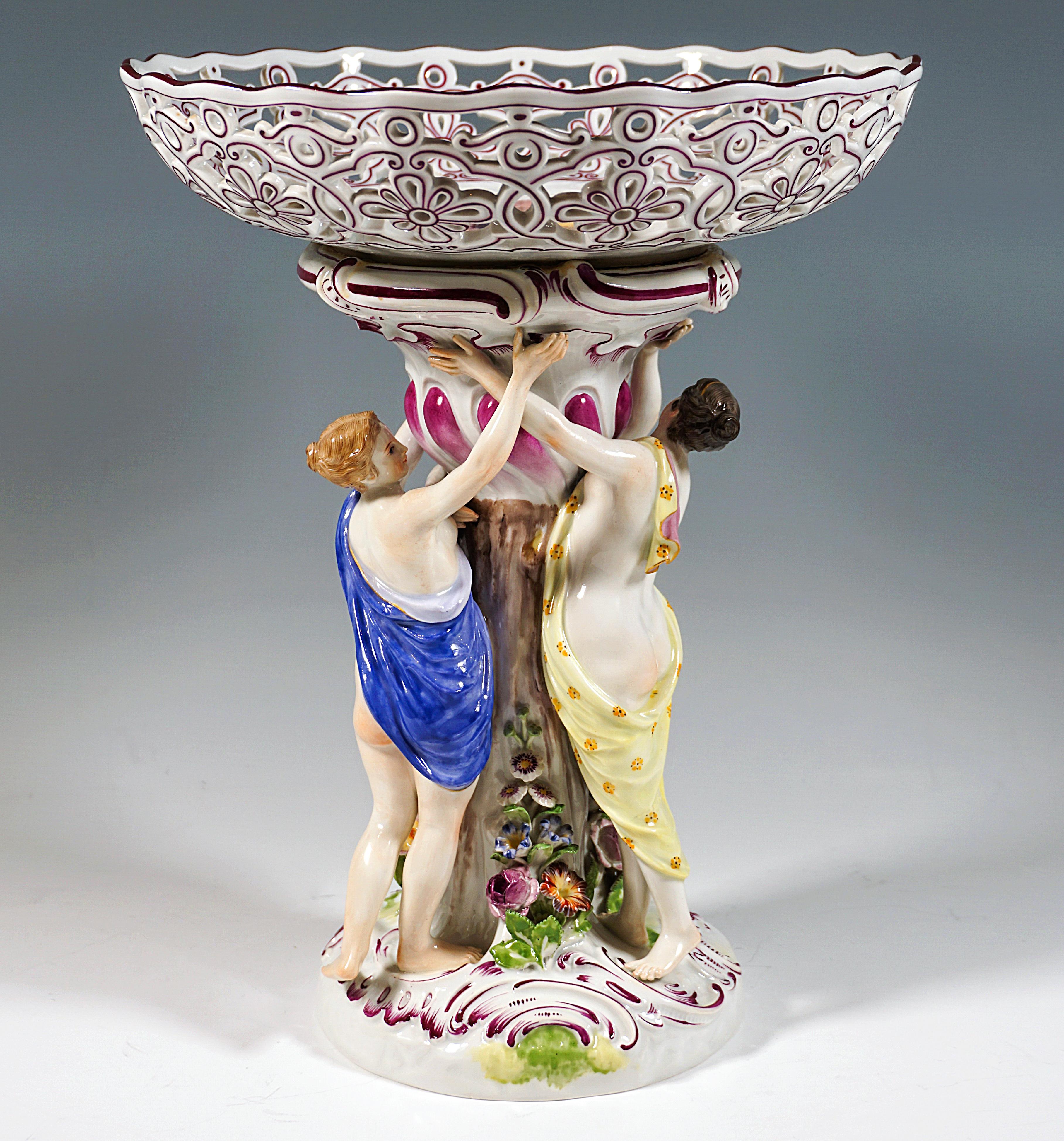 Meissen Piece with excellent fruit painting:
On a round, low rock base with purple heightened rocailles in the center a tree trunk, on which from the ground vividly formed floral bunches are climbing up, three young graces wrapped only in cloths
