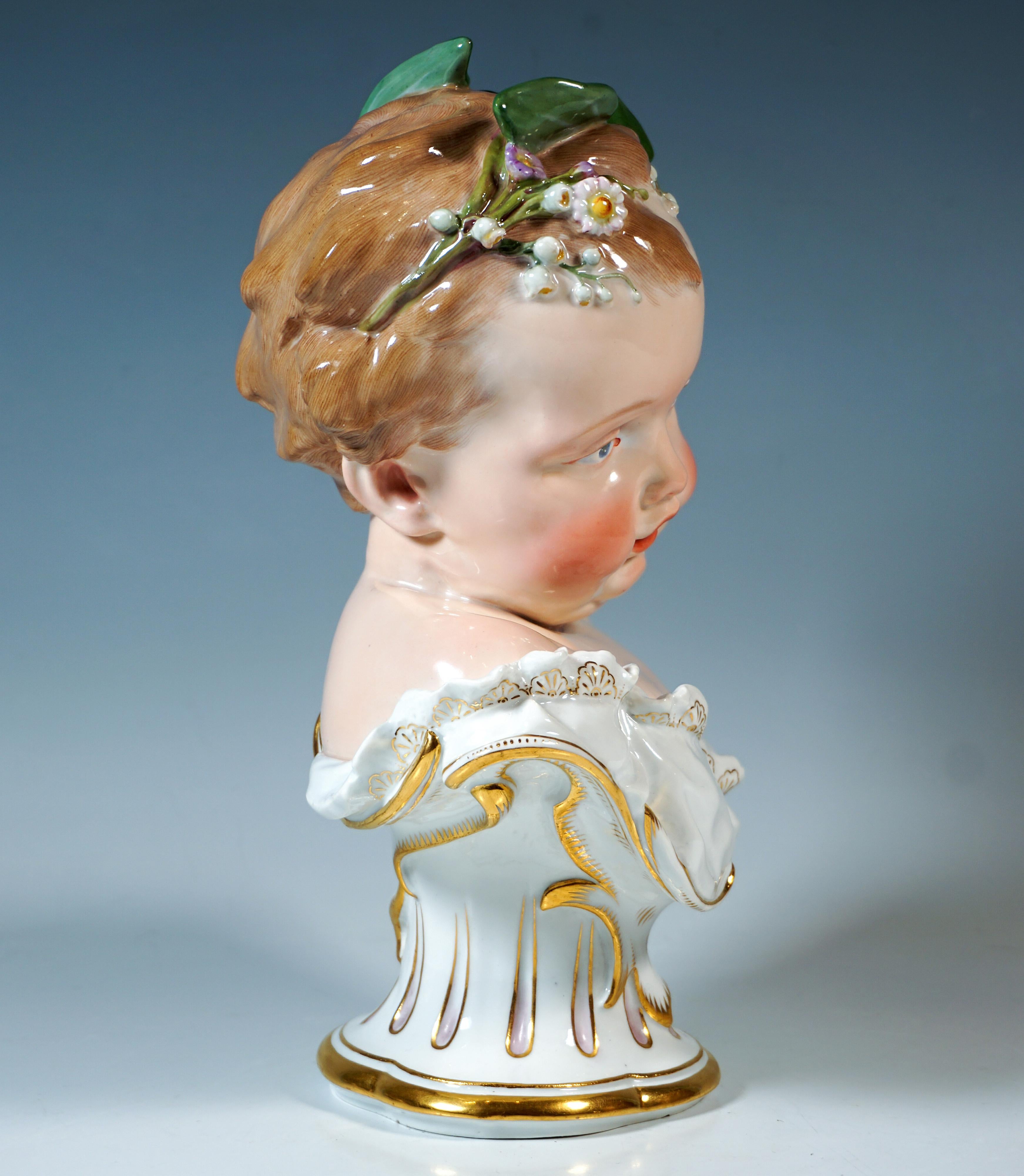 Sehr seltene Büste eines in weißes Tuch gehüllten Engels mit Goldverzierung, dessen blonde Locken mit zarten Frühlingsblumen und Blättern geschmückt sind und dessen Kopf leicht zur Seite geneigt ist.
Gewölbter Sockel mit Vierblattmuster, Rocaille