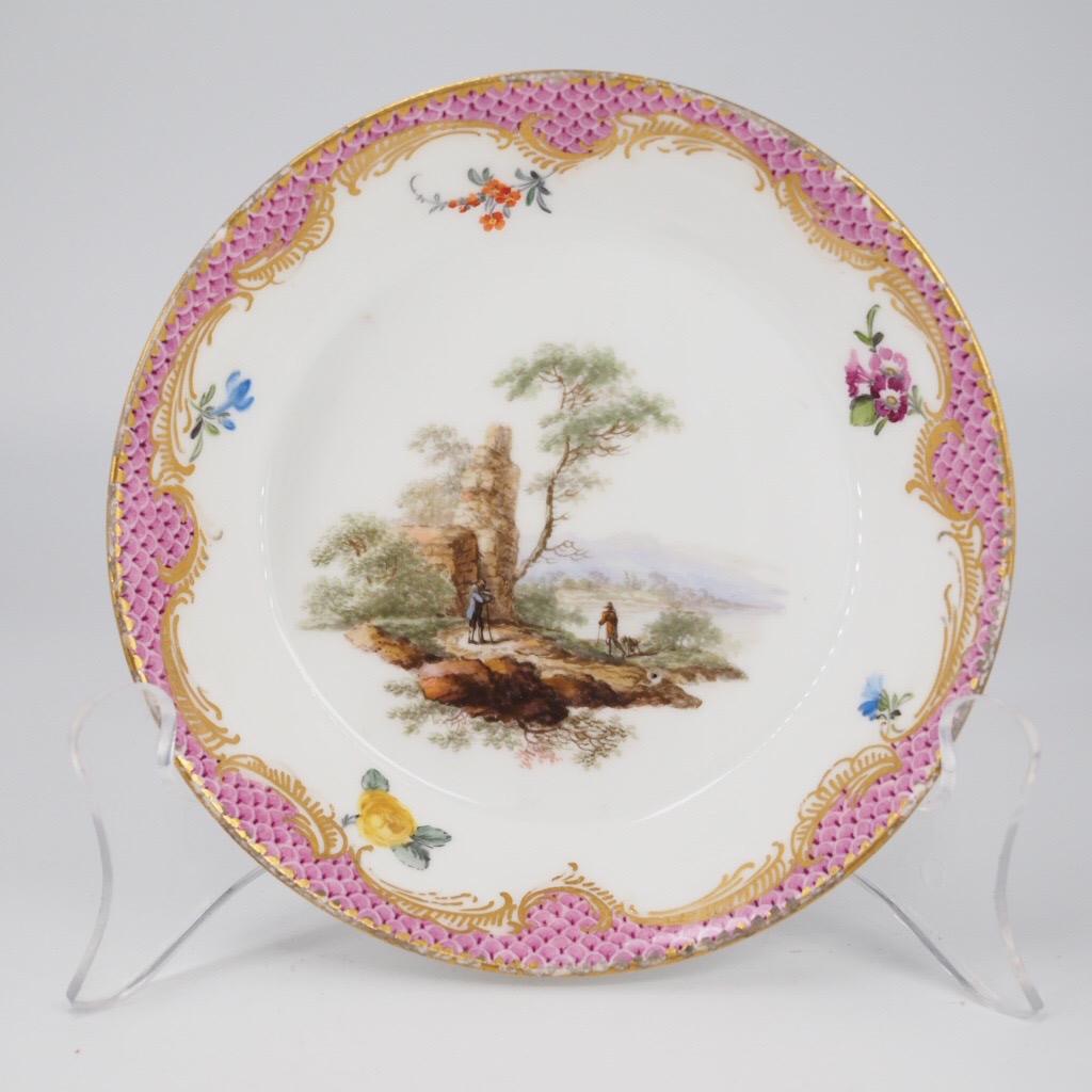 Meissen coffee cup & saucer, the handle an elegant double twig with acanthus leaf terminals, well painted with scenes of men in landscapes, within a purple scale border with Rococo gilt scroll edging, within a dentil gold rim. 

Blue crossed
