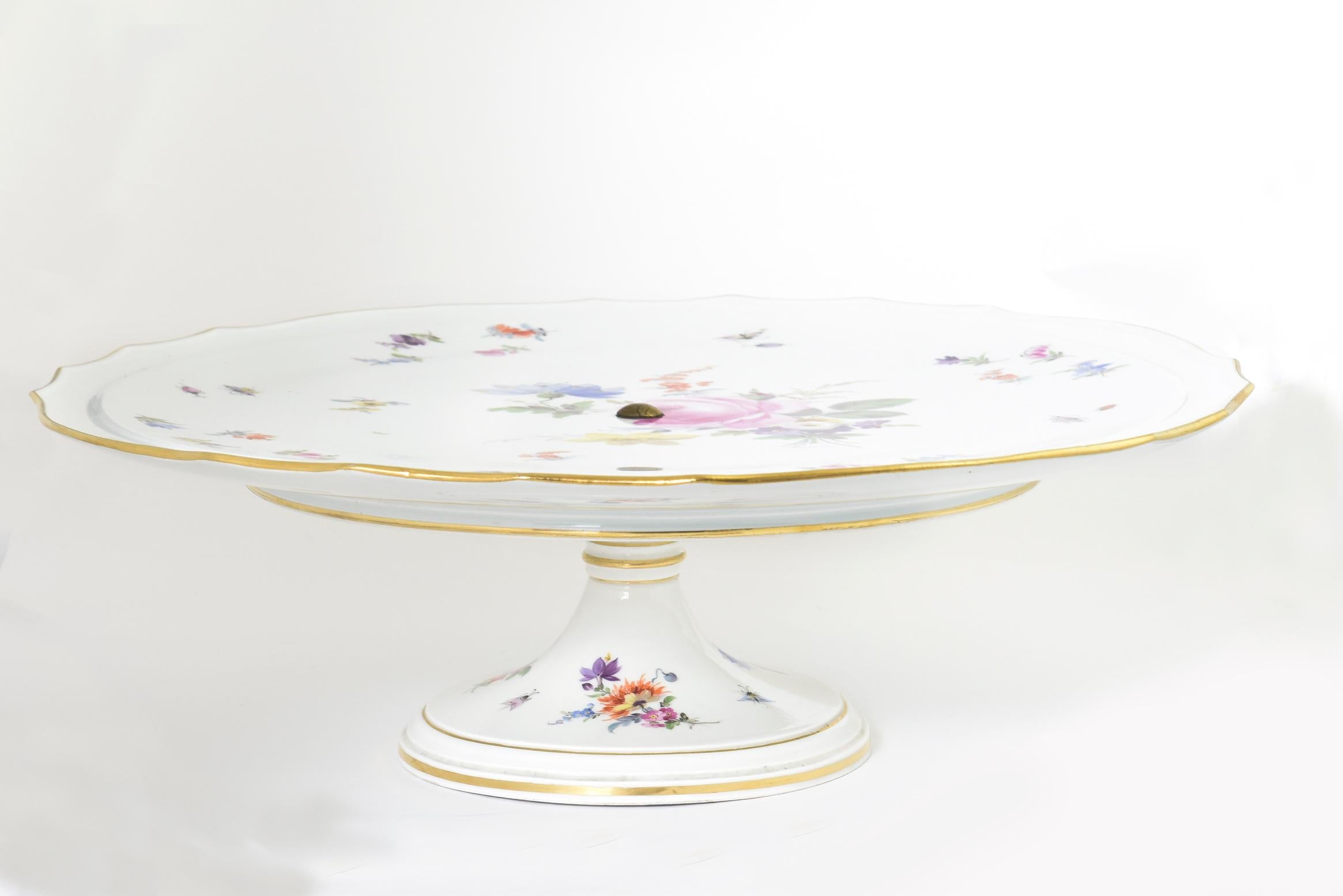 A Meissen cake Stand with a gilt scalloped rim decorated with hand painted scattered flowers, bugs, and butterflies. Cross swords in blue under glaze, impressed,
circa first half of the 20th century.