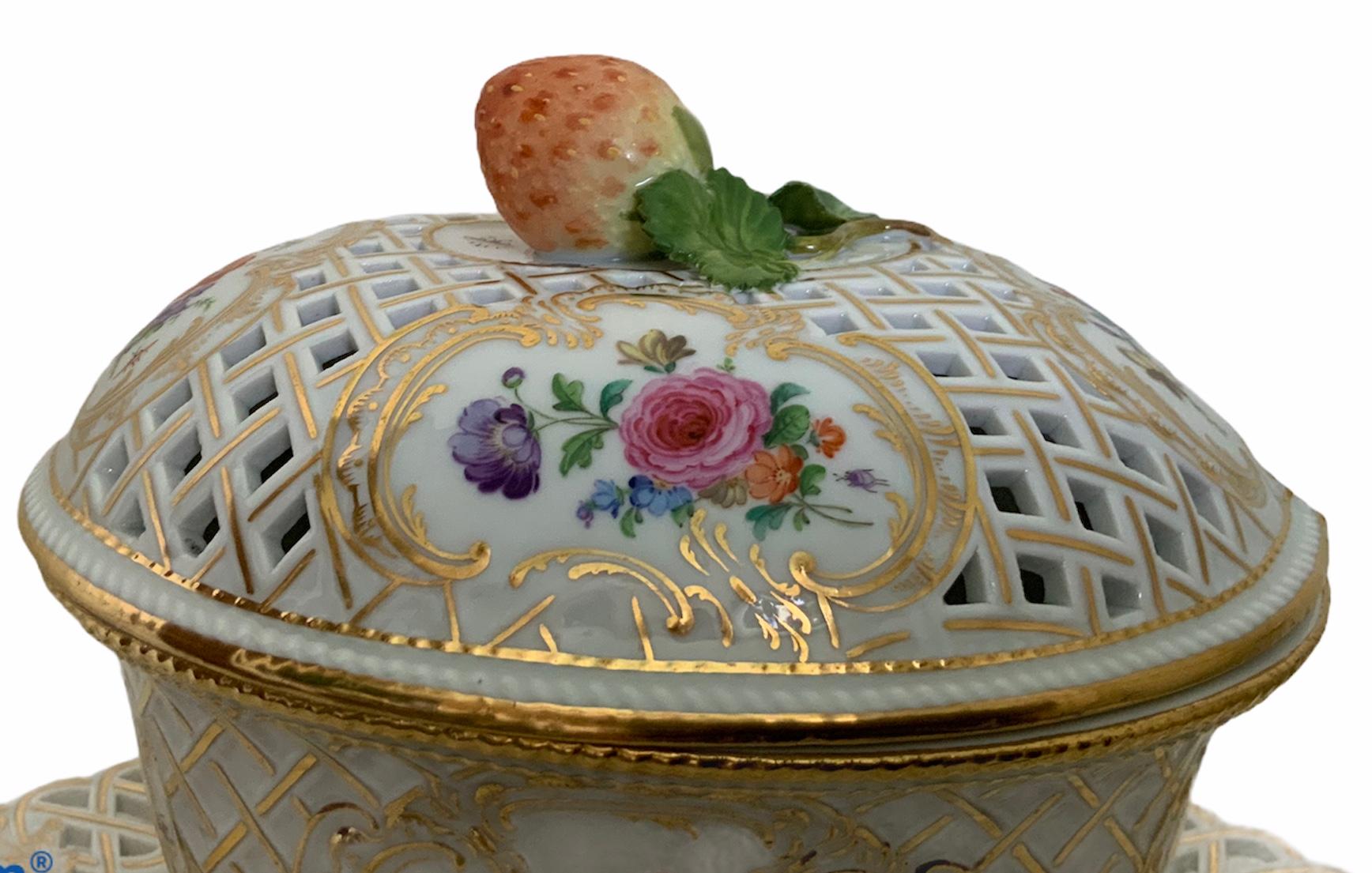 This a Meissen hand painted porcelain gravy/sauce oval tureen with attached diamond shaped plate. The lid has reticulated gilt braided pattern and adorned with four gilt rocaille scrolled border cartouches that have different flowers in the center.