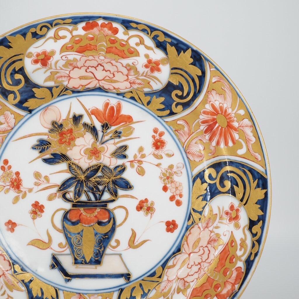 Meissen plate decorated in rich Imari colours after a Japanese original, the center painted in underglaze blue, iron red and gold with a vase of flowers standing on a low table, the border with four panels of a moth and peony within a shaped reserve