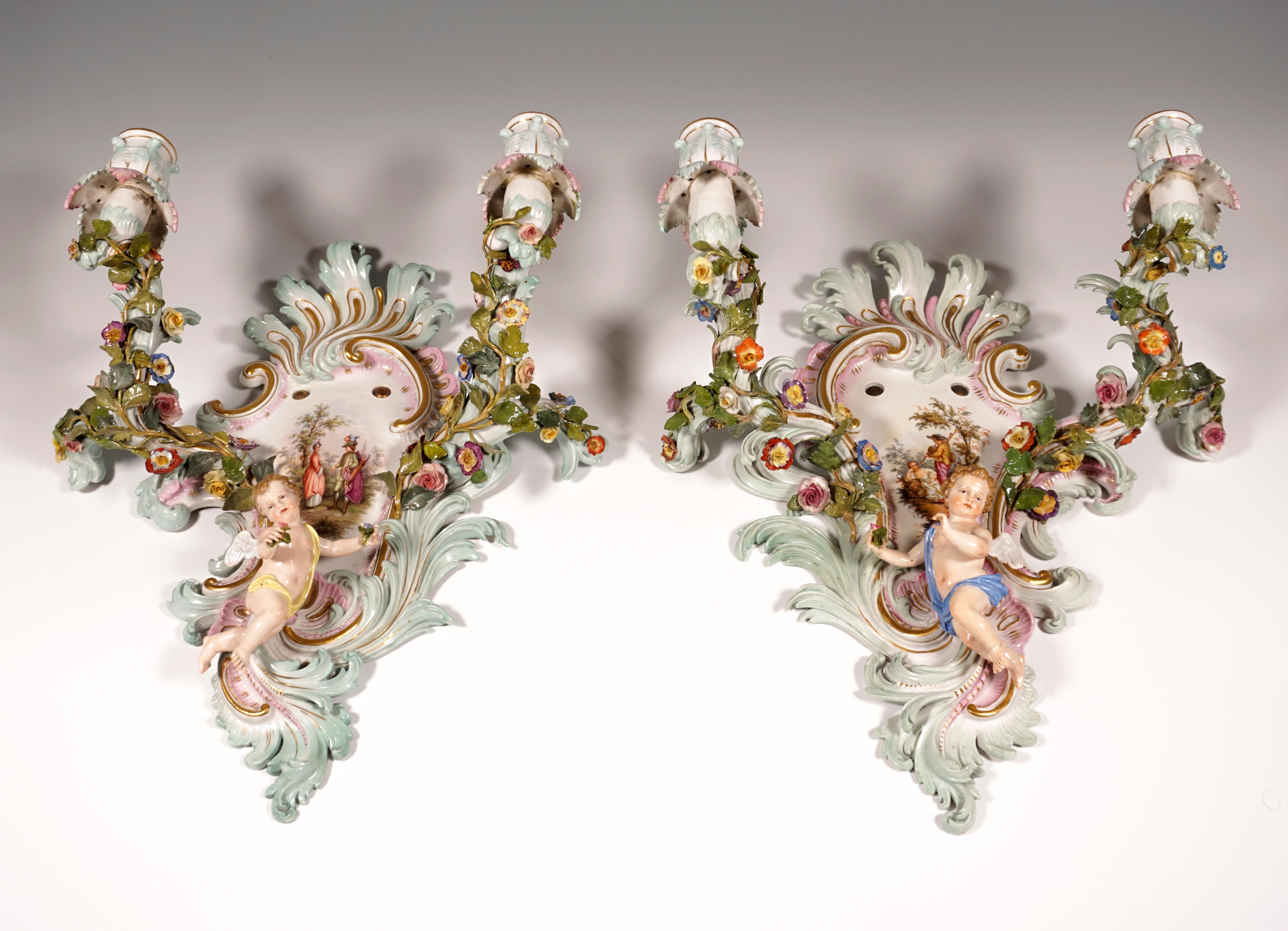Pair of two-flame wall appliqués in the form of colored and gold-decorated rocaille shields, each with two candlestick arms richly covered with flowers and leaf tendrils, as well as one floating, winged cupid boy holding flowers in his hands, in the