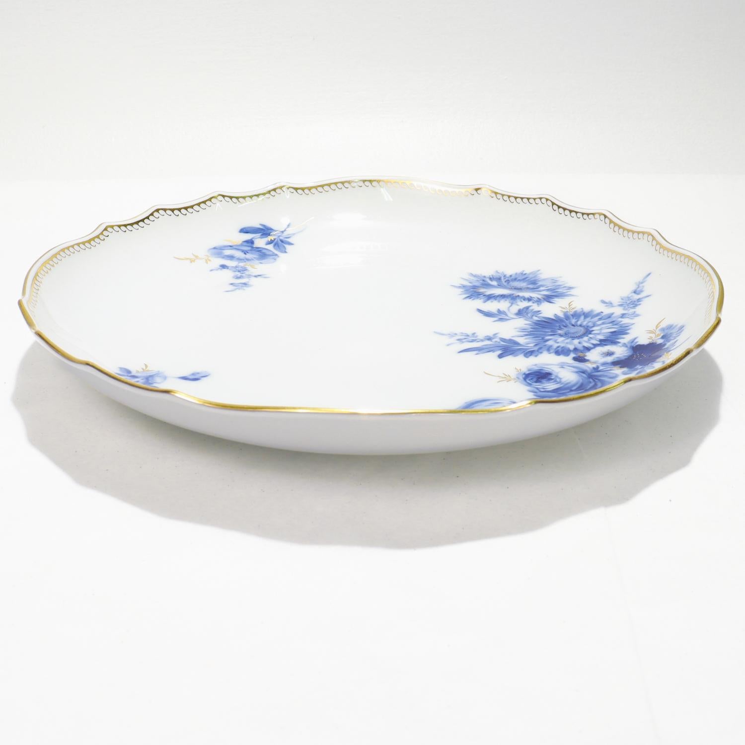 A fine antique porcelain serving dish or large plate.

By the Meissen Porcelain Manufactory.

Decorated with blue underglaze flowers with gilt stems and twigs on a white ground.

The scalloped rim with gilding around its circumference and a gilt