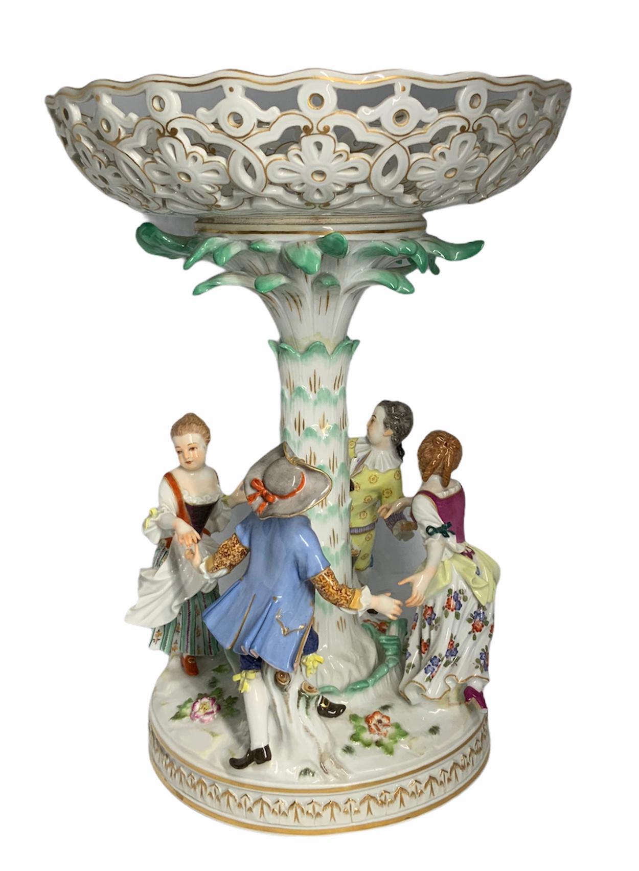 This is a Meissen porcelain centerpiece depicting a group of children holding hands & dancing around a tall palm tree. This serves as a pedestal base to the decorated and reticulated wide compote that is embellished with flowers & scrolls. The