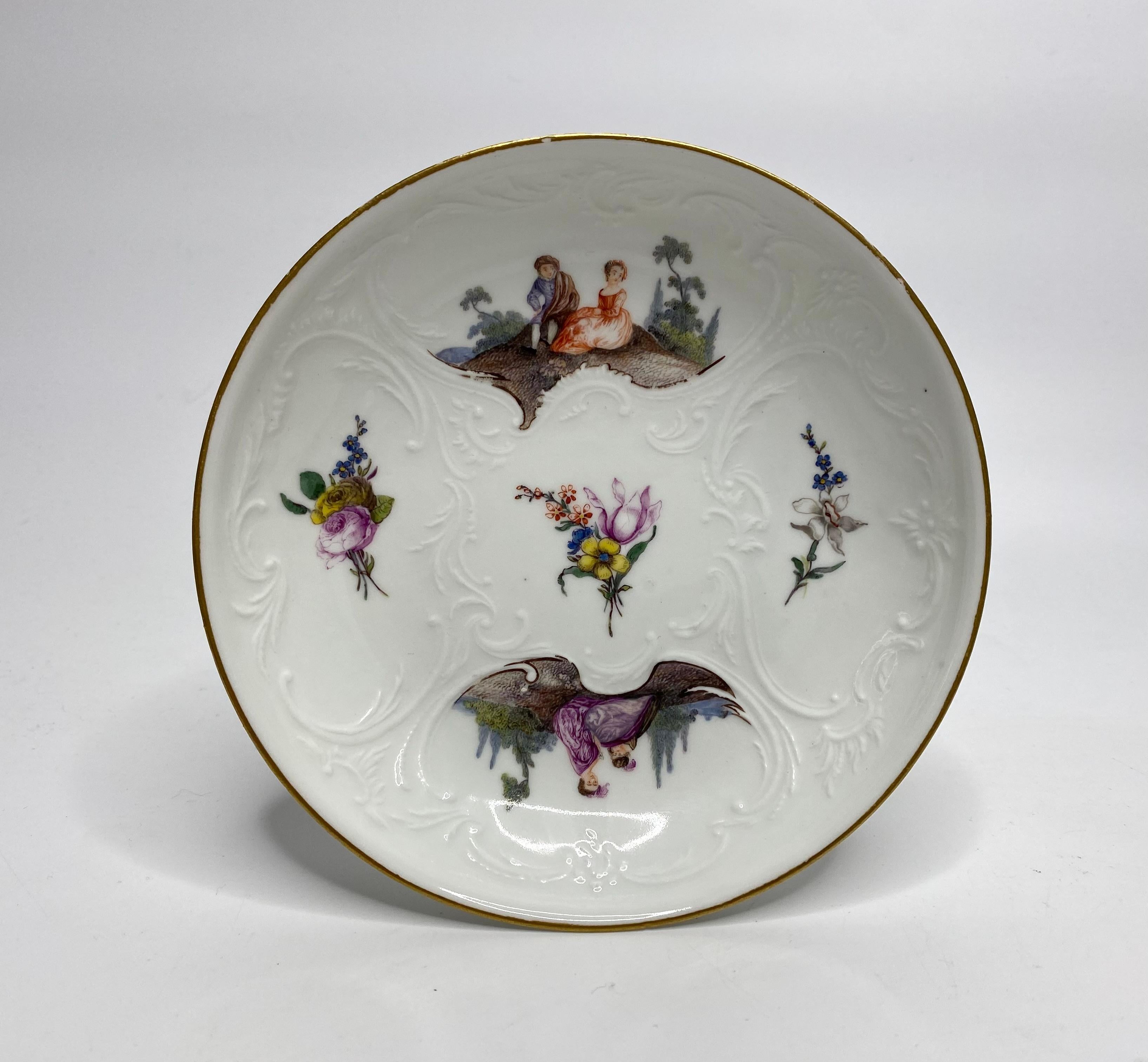 Meissen porcelain cup and saucer, c. 1740. Both the cup and the saucer, finely moulded in blind relief, with scrollwork panels. The panels painted in enamels, with scenes of courting couples in 18th Century attire, in the countryside; one couple