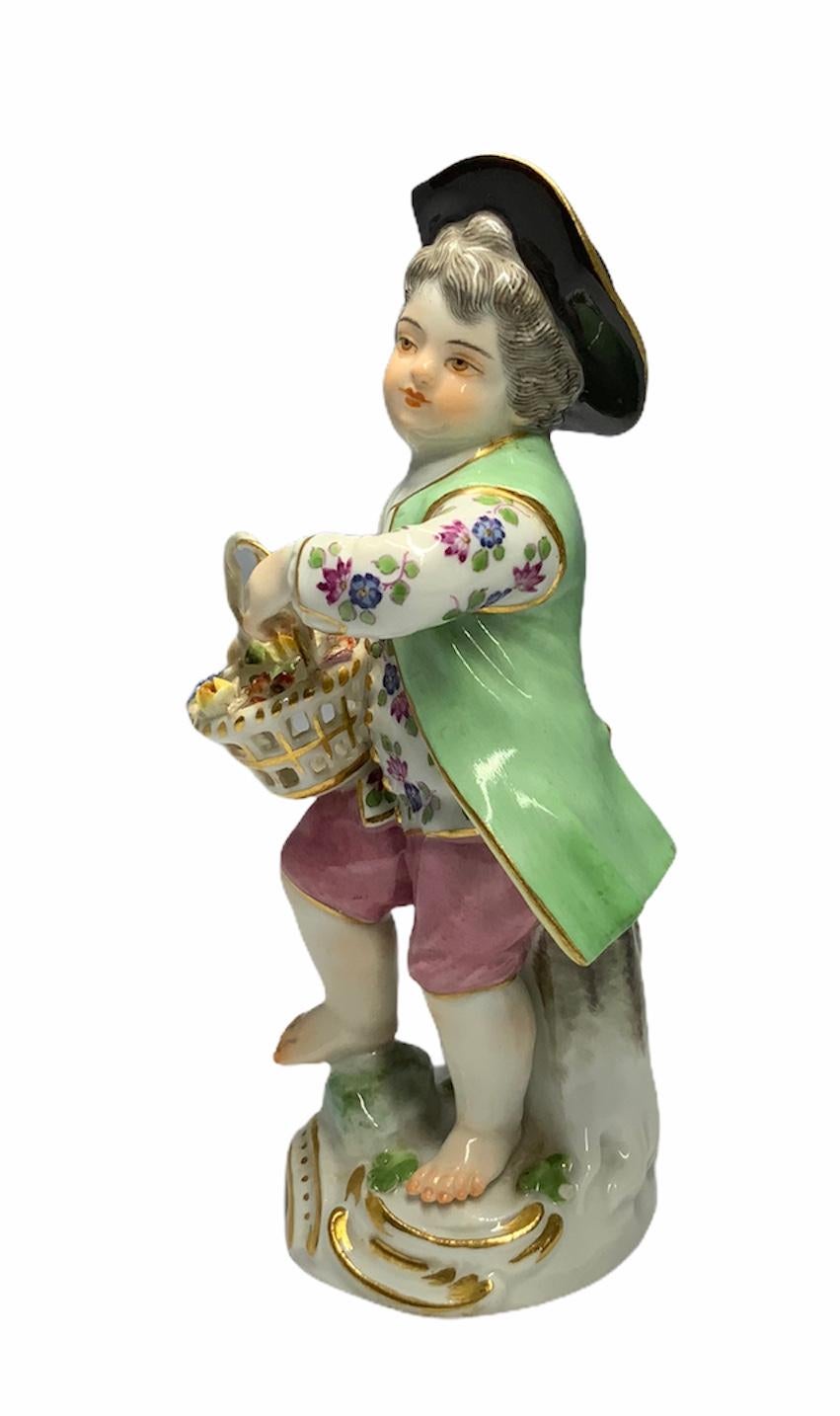 This a Meissen porcelain figure depicting a boy dressed with colorful clothes and wearing a black hat. He appears to be gathering flowers in a basket. He is standing in front of a tree trunk. Under a gilt white rocaille base appears the Meissen