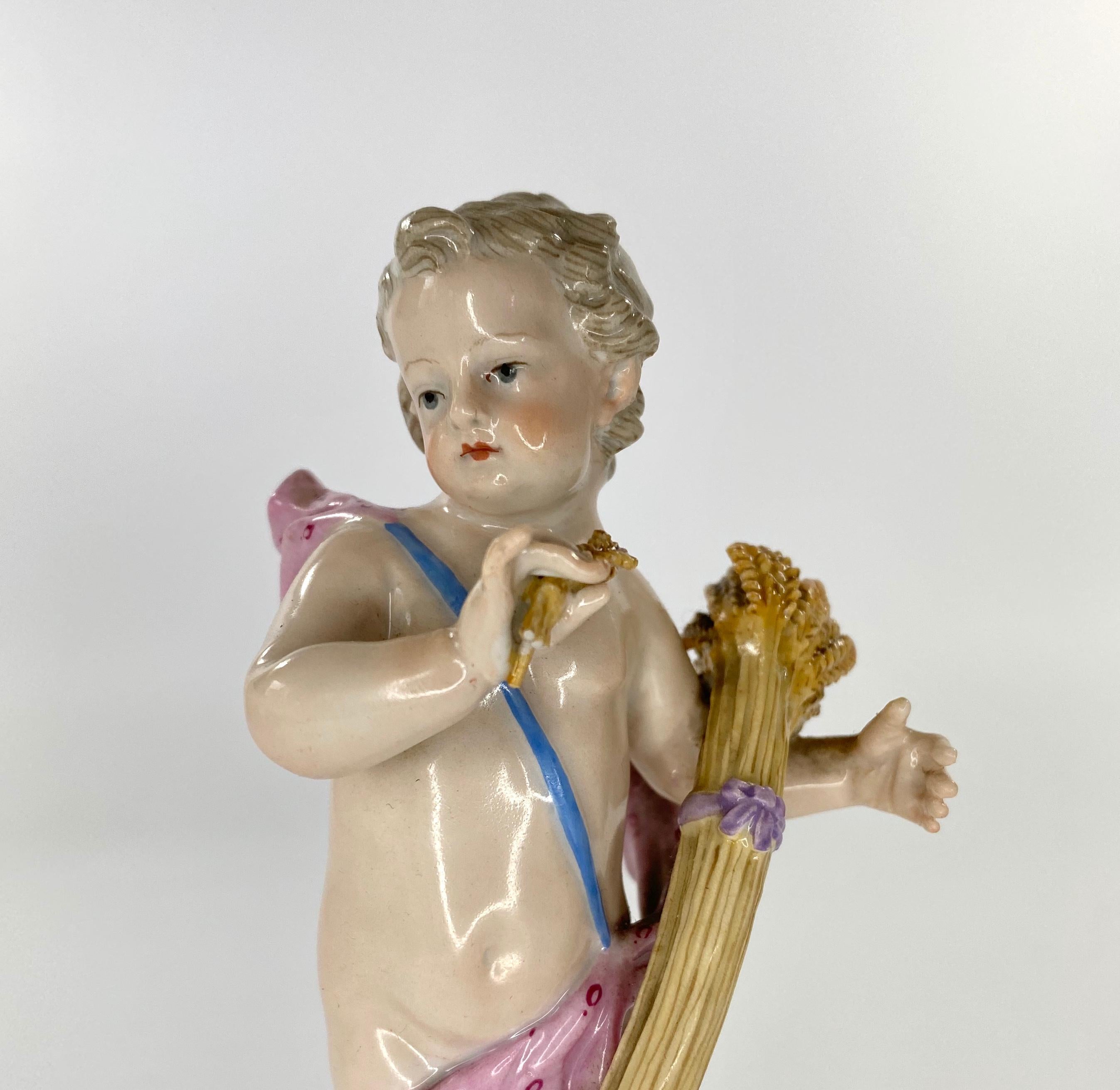 Meissen porcelain Putti, c. 1870. The standing Putti, representing ‘Autumn’ from The Four Seasons’, holding a large sheaf of corn in his left arm, whilst holding a smaller sheaf in his right hand. Set upon a circular grassy mound, with a gilt
