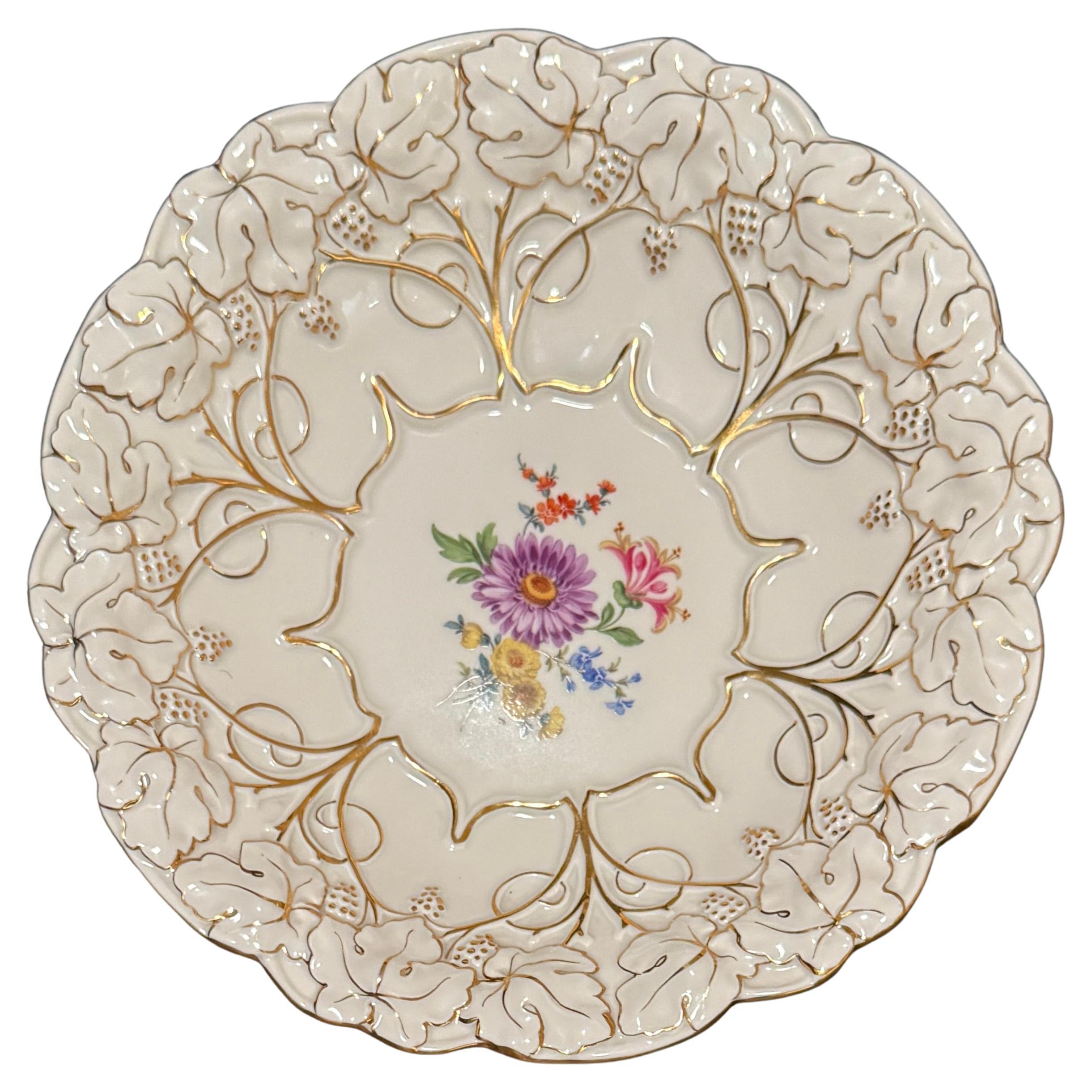 Superbe exemple du savoir-faire de Meissen, cette assiette en porcelaine peinte à la main présente une bordure finement réticulée de feuilles de vigne et de vignes en relief, rehaussée d'une dorure appliquée à la main. Le bord ondulé et le motif