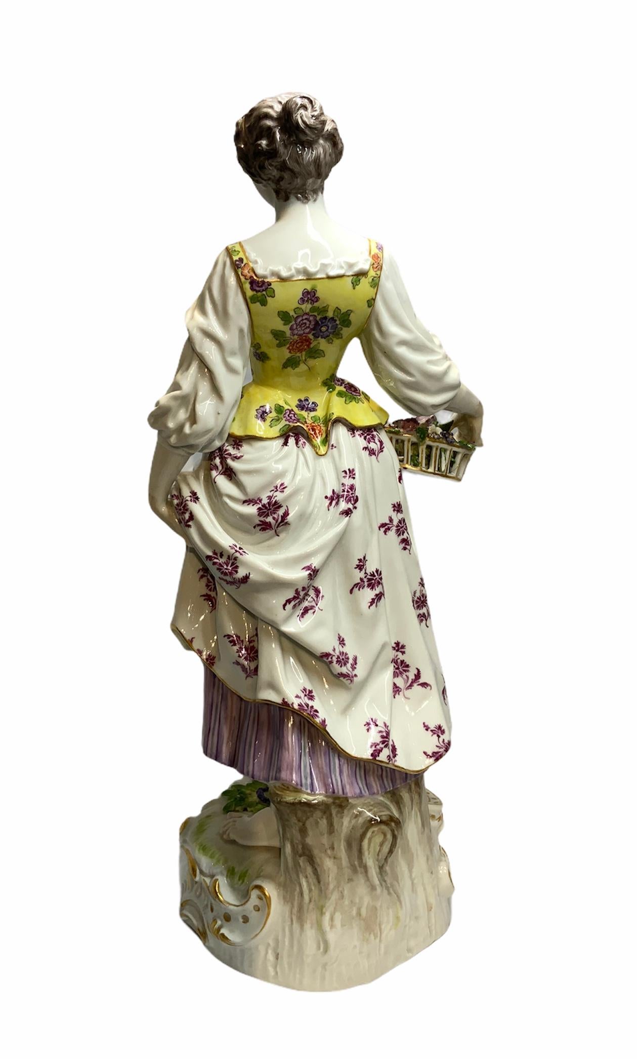This is a Meissen porcelain depicting a young shepherdess dressed with a colorful flower’s fabric. She is barefooted over a gilt rocaille white base and standing in front of a tree trunk. She is also holding a basket of flowers in her right hand.