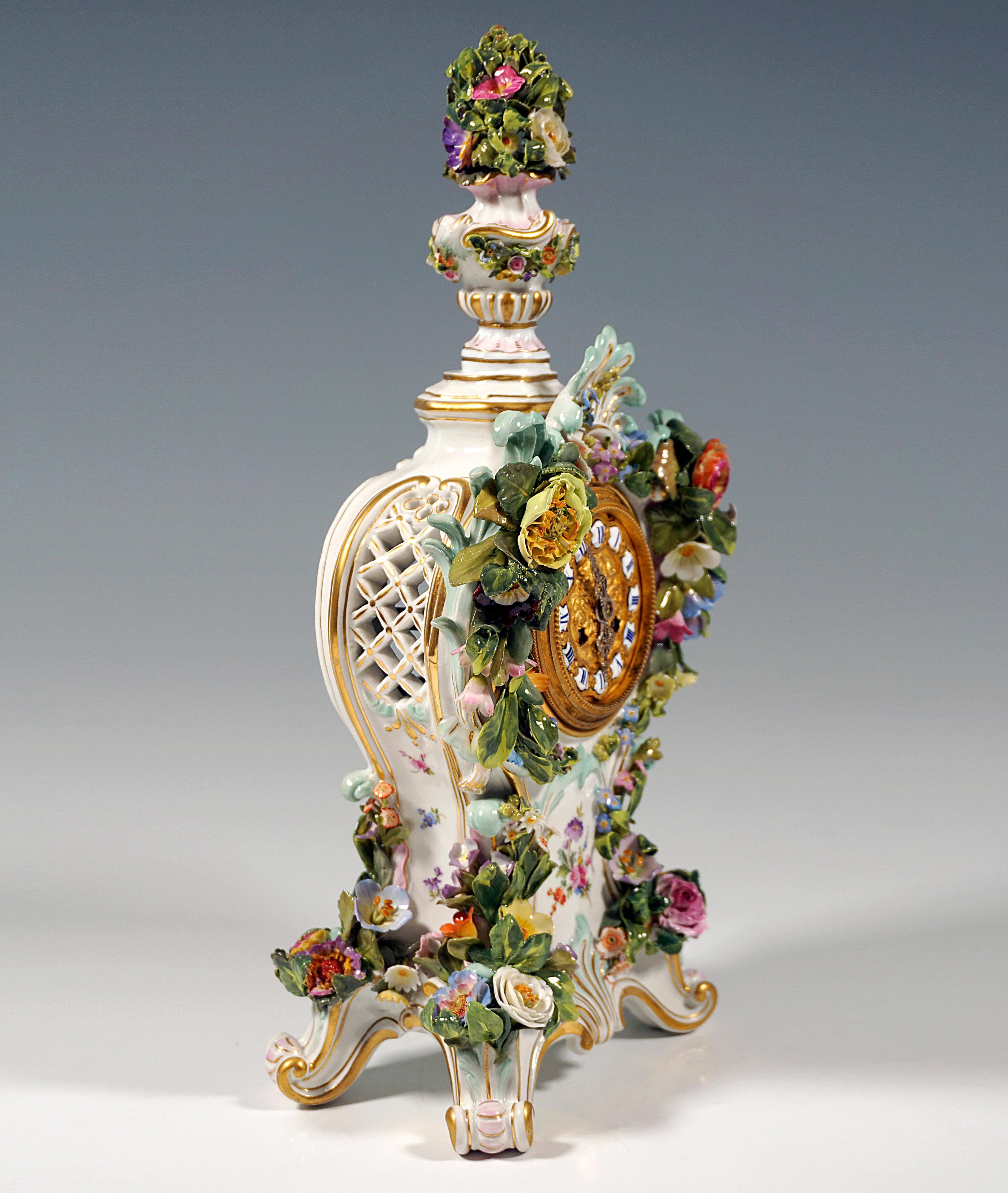 Clock case rising on four gold heightened rocaille feet, richly decorated with delicate, vividly formed flowers, leaves and rocaille surrounding the dial, laterally pierced like a lattice, crowned at the top by a colourfully decorated and gold